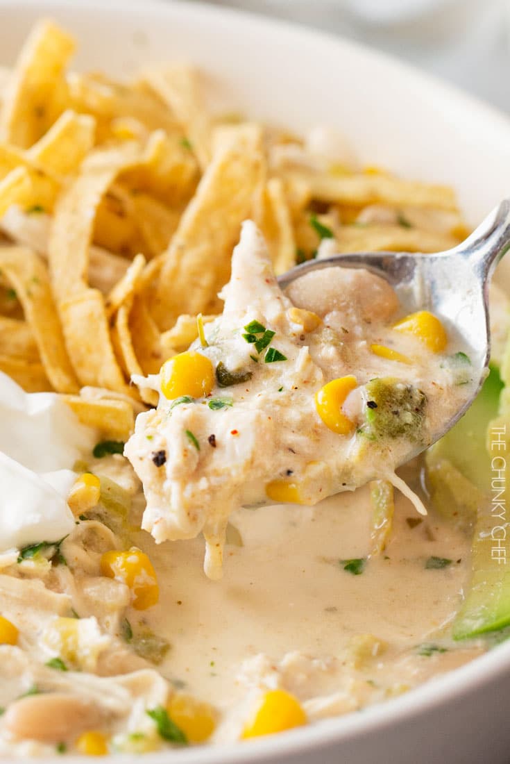 A spoonful of creamy chicken chili with corn, garnished with herbs, next to tortilla strips and a dollop of sour cream in a bowl—perfect for fans of high protein recipes and hearty crockpot recipes.