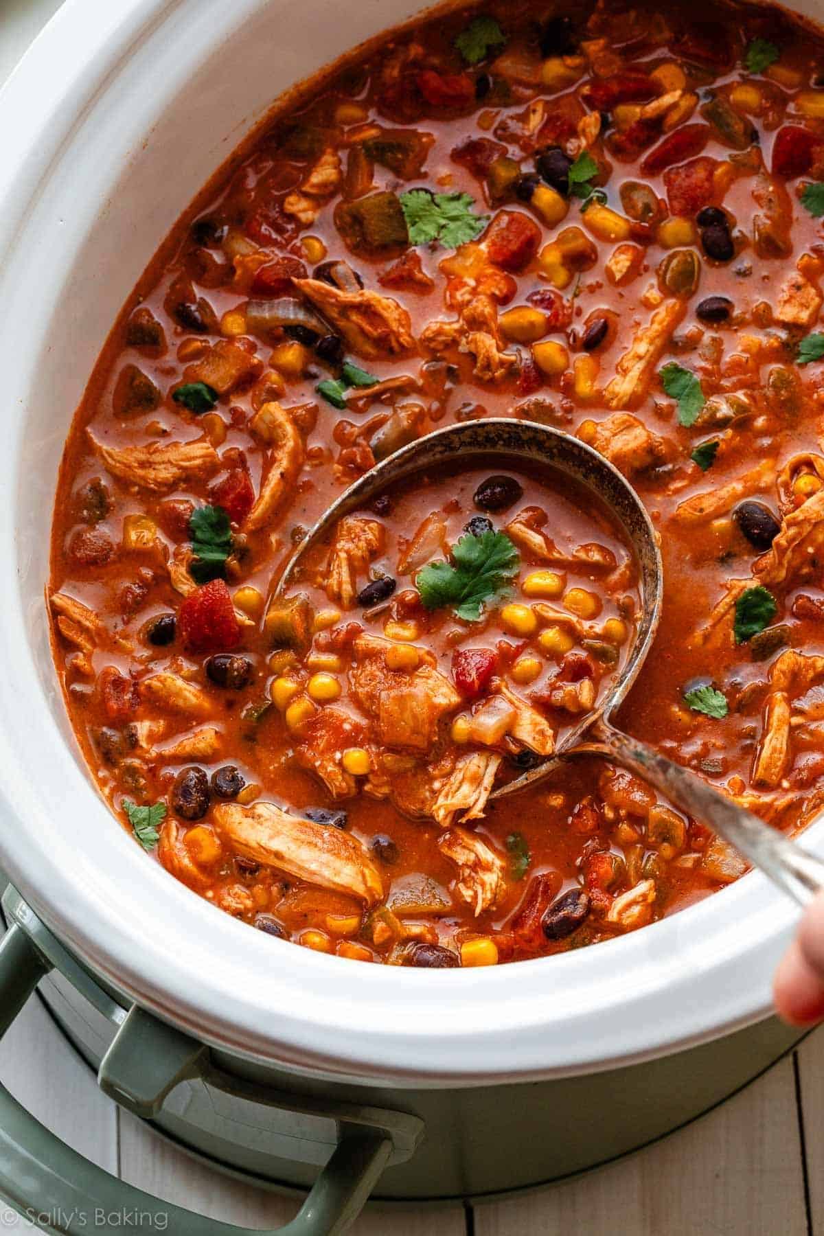 A ladle scoops hearty, high protein chicken chili with beans, corn, tomatoes, and cilantro from a slow cooker—perfect for fans of easy crockpot recipes.