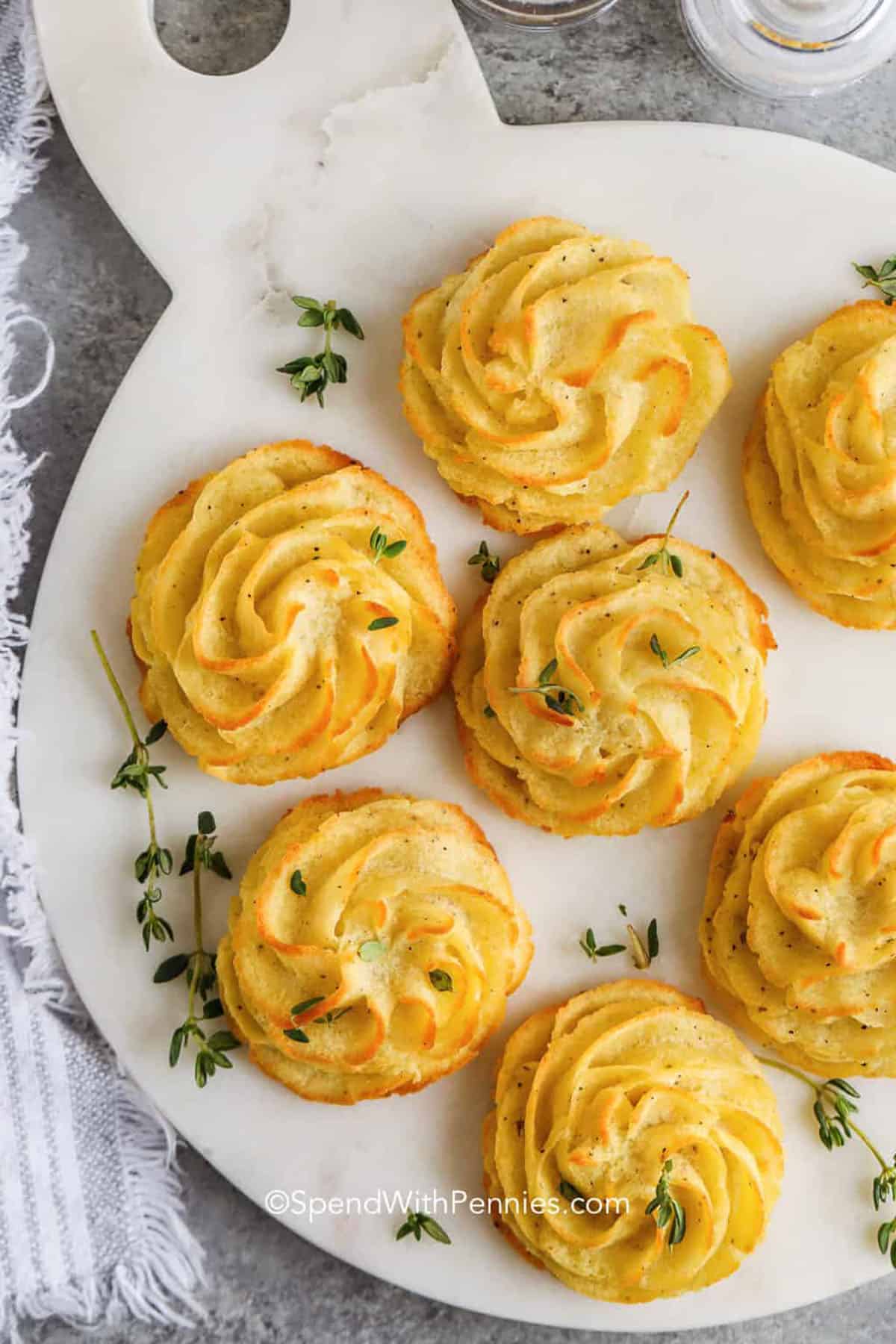 Six piped swirls of duchess potatoes garnished with fresh herbs are arranged on a white serving board—an elegant touch for your Easter party food spread.
