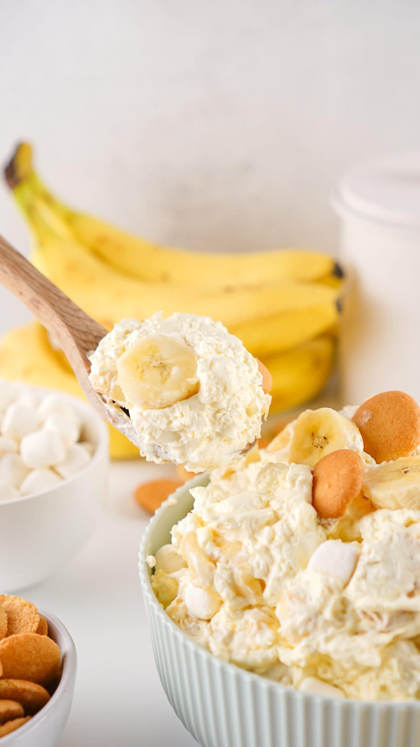 A bowl of banana pudding topped with vanilla wafers and banana slices; a spoon holds a serving. Bananas and a bowl of marshmallows are in the background, making this a sweet choice for Easter side dishes.