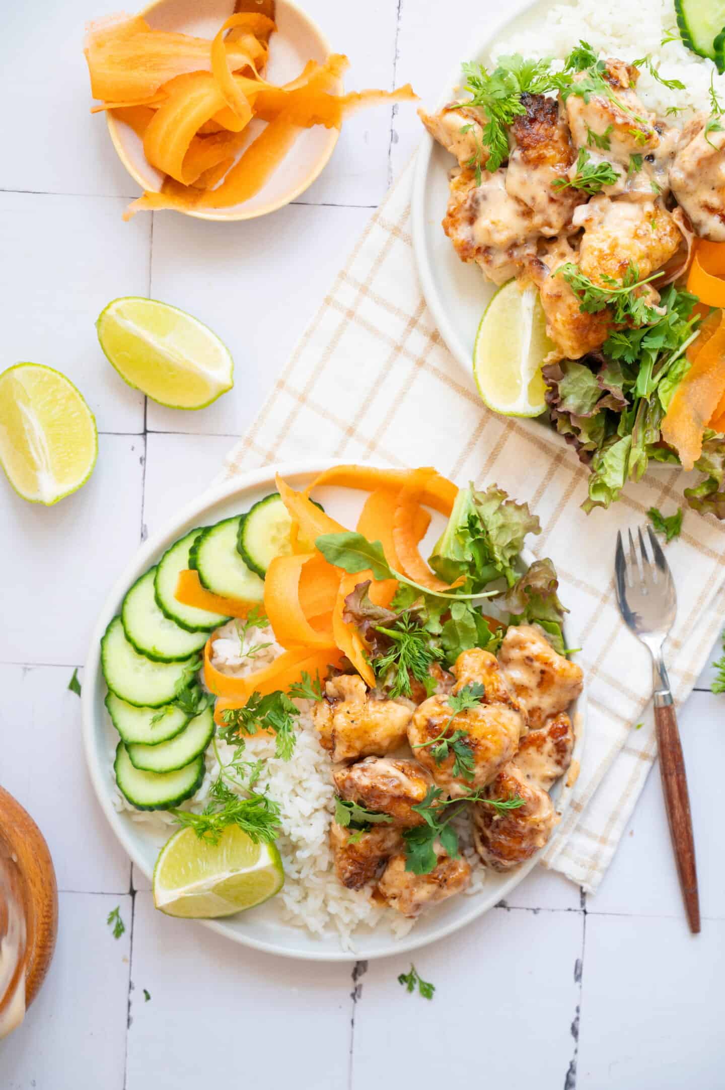 A vibrant Chicken Rice bowl featuring sauced chicken, sliced cucumbers, leafy greens, carrot ribbons, fresh herbs, and a lime wedge atop fluffy rice. Served with a fork and extra carrot ribbons on the side for a classic Bang Bang Chicken touch.