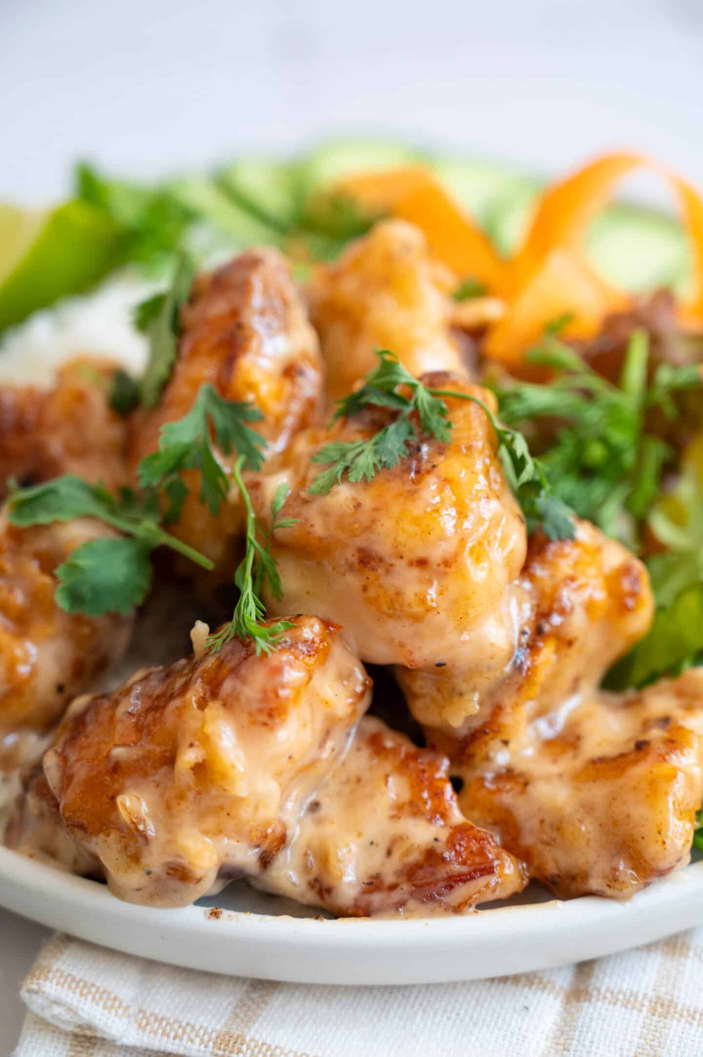 A close-up of creamy, glazed Bang Bang Chicken pieces garnished with fresh cilantro, served alongside sliced cucumbers and carrots on a white plate.