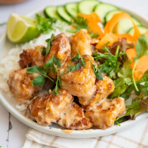 A Bang Bang Chicken Rice Bowl featuring sauced chicken pieces served with white rice, cucumber slices, carrot ribbons, mixed greens, and a lime wedge.