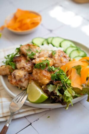 Plate of Bang Bang Chicken with creamy sauce, garnished with parsley, served alongside sliced cucumber, mixed greens, carrot ribbons, a lime wedge, and a fork on the side.