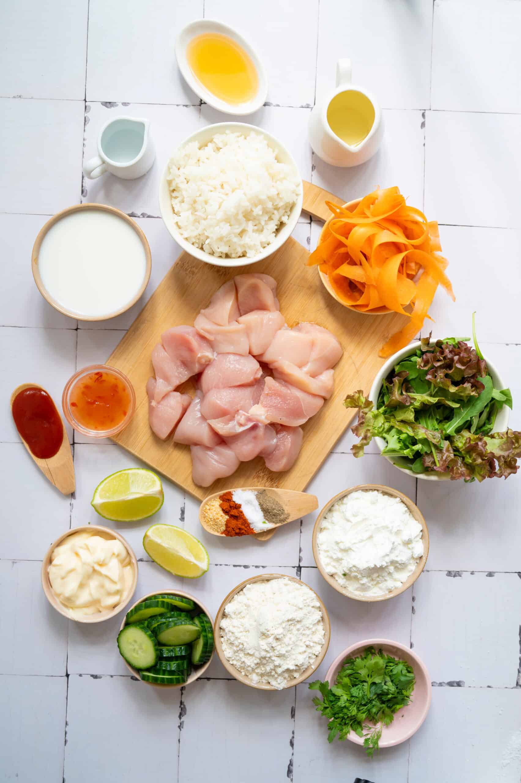 Various fresh ingredients arranged on a white surface, including raw chicken pieces ideal for Chicken Rice, rice, leafy greens, sliced carrots, cucumber, sauces, spices, flour, and liquids in bowls and pitchers.