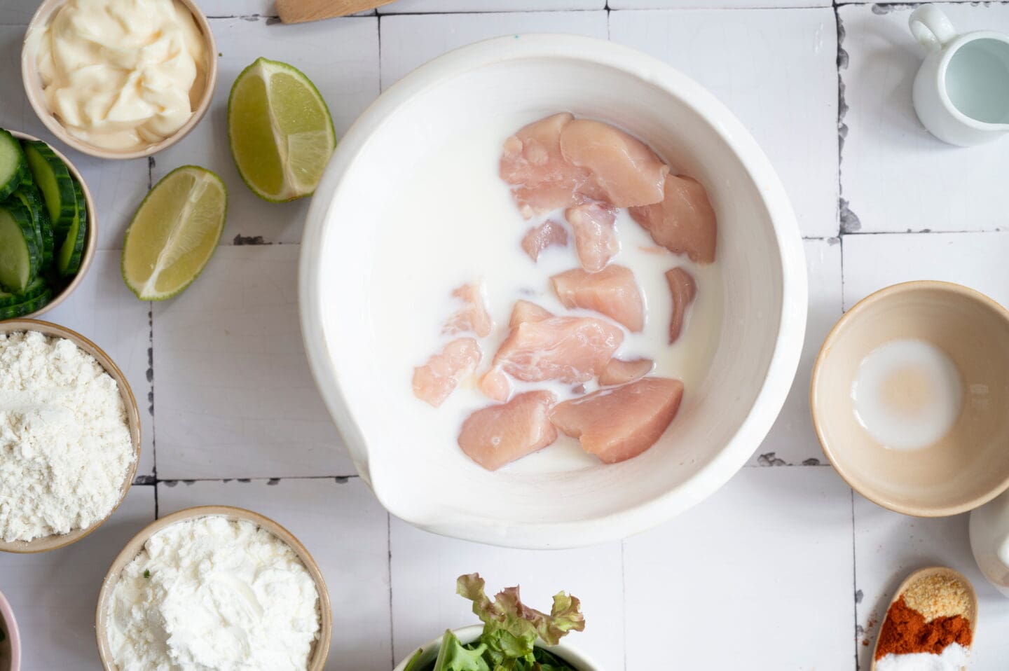 Sliced raw chicken pieces soaking in a bowl of milk, surrounded by various ingredients for a flavorful Bang Bang Chicken Rice Bowl, all arranged on a white tiled surface.