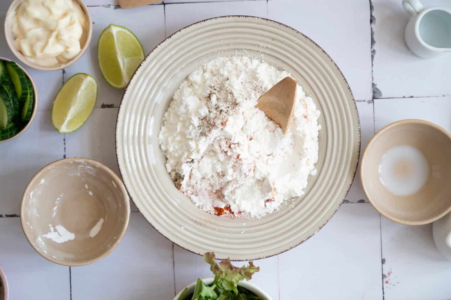 A bowl with flour and seasonings being mixed for Bang Bang Chicken, surrounded by lime wedges, empty bowls, and other ingredients on a white tiled surface.
