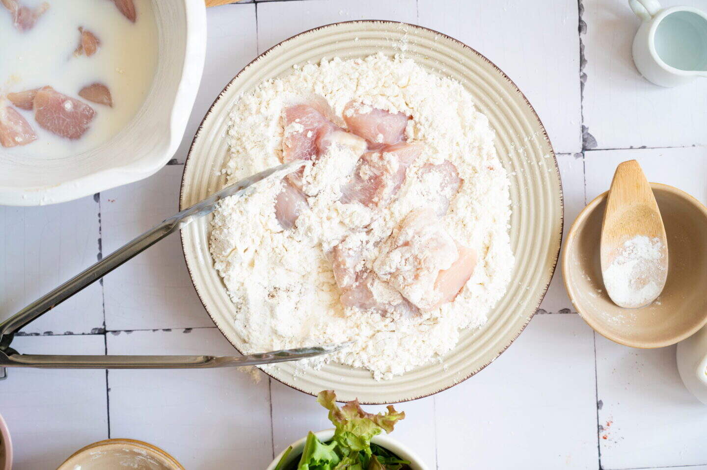 Raw chicken pieces being coated in flour on a plate, with tongs nearby and various bowls of ingredients—perfect prep for a flavorful Bang Bang Chicken or delicious Chicken Rice.