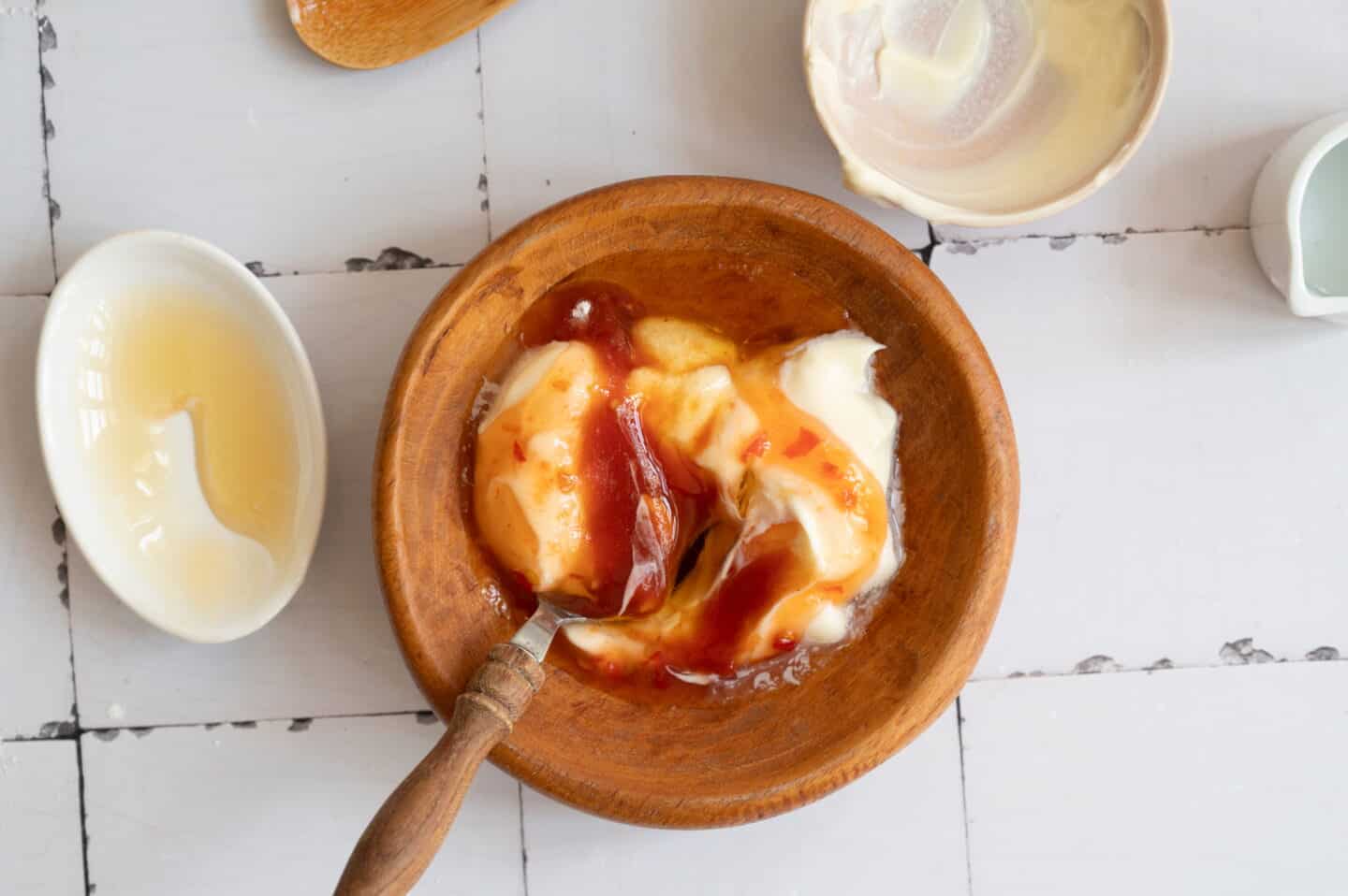 A wooden bowl with mayonnaise, ketchup, and mustard being mixed with a spoon—perfect for drizzling over a Chicken Rice Bowl—surrounded by small dishes with condiments on a white tiled surface.