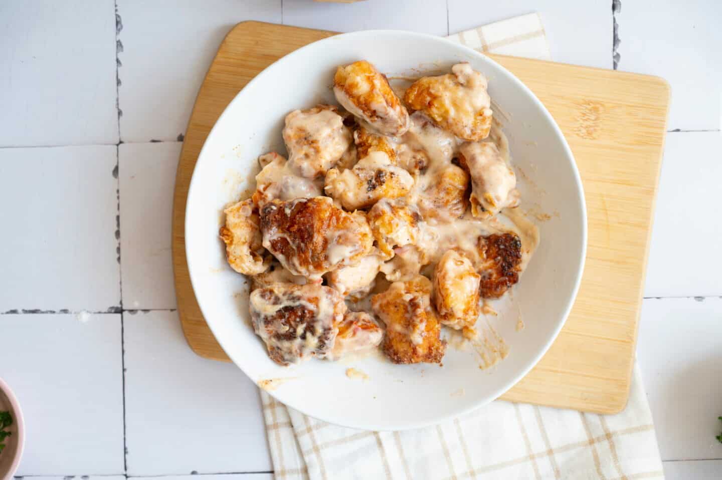 A white bowl filled with Bang Bang Chicken pieces coated in a creamy sauce, placed on a wooden cutting board atop a tiled surface.