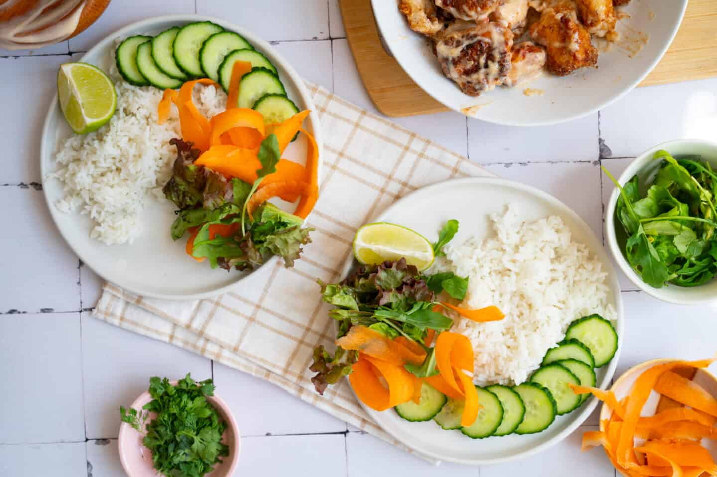 Two plates of Bang Bang Chicken Rice Bowl with white rice, sliced cucumber, carrot ribbons, mixed greens, and lime wedges on a cloth, surrounded by herbs, salad, and a dish of flavorful cooked chicken.