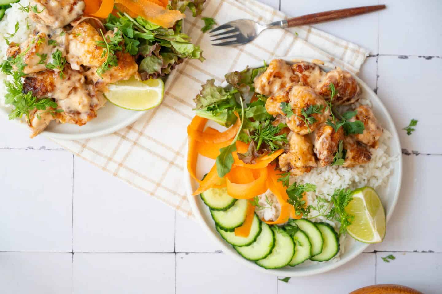 A vibrant Chicken Rice bowl featuring sauced chicken, sliced cucumber, carrot ribbons, fresh greens, and a lime wedge, served on a table with a fork and another plate partially visible.