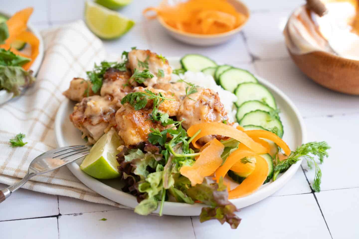 A plate of Bang Bang Chicken with creamy sauce, sliced cucumbers, mixed greens, carrot ribbons, and a lime wedge, served on a white plate with a fork.