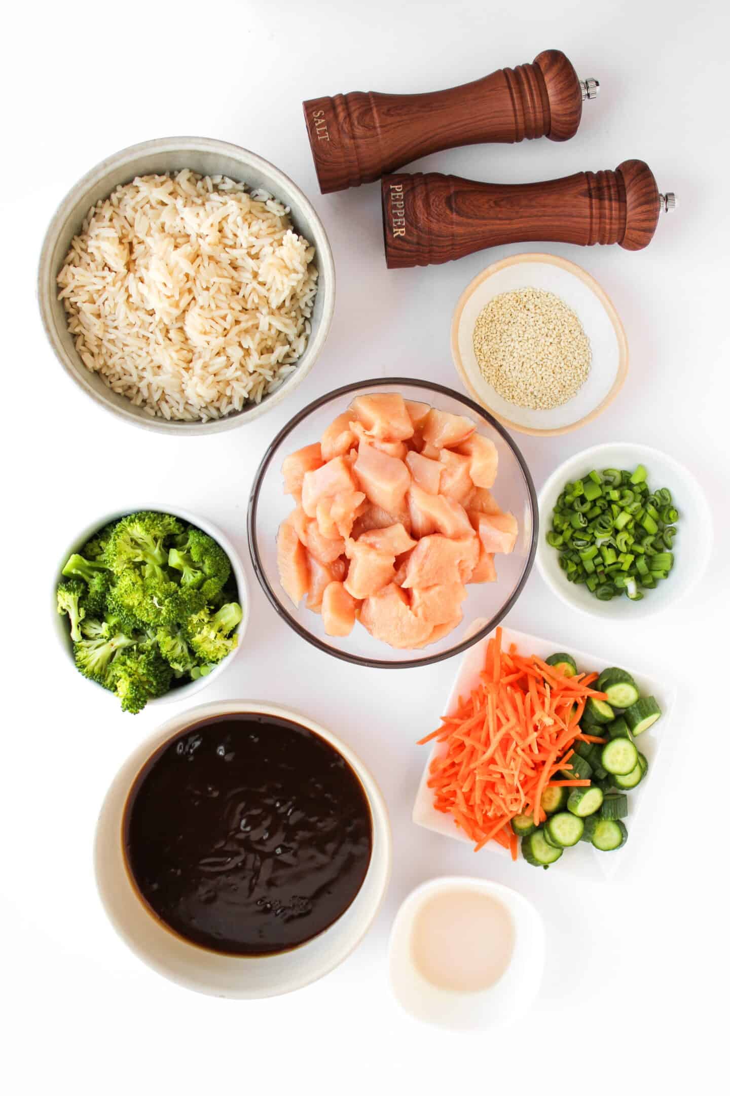 Overhead view of bowls with ingredients for Teriyaki Chicken Bowls: rice, diced raw chicken, broccoli, sliced carrots, cucumber, green onions, sauce, oil, sesame seeds, and two pepper grinders on a white surface.