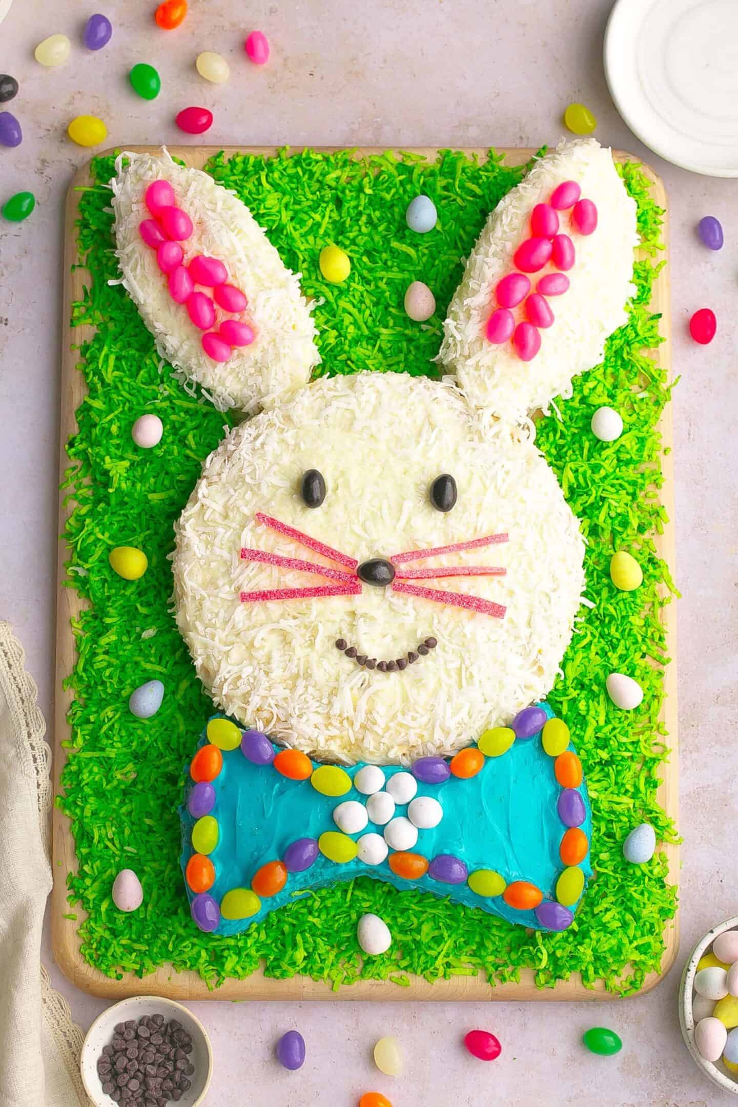 A rectangular cake decorated to look like a bunny with white icing, pink and colored candies, green shredded coconut grass, and a blue bow tie, surrounded by candy eggs—perfect Easter party food for your celebration.