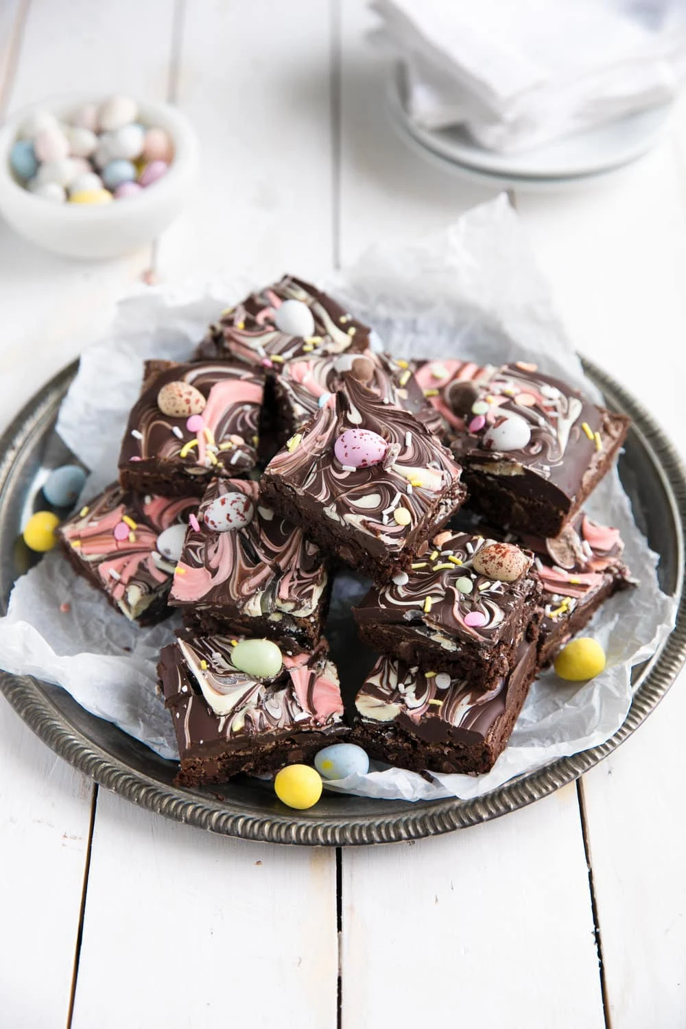 A plate of chocolate brownies topped with pink and white marbled icing and mini candy eggs, arranged on parchment paper—perfect for Easter recipes or festive Easter party food.