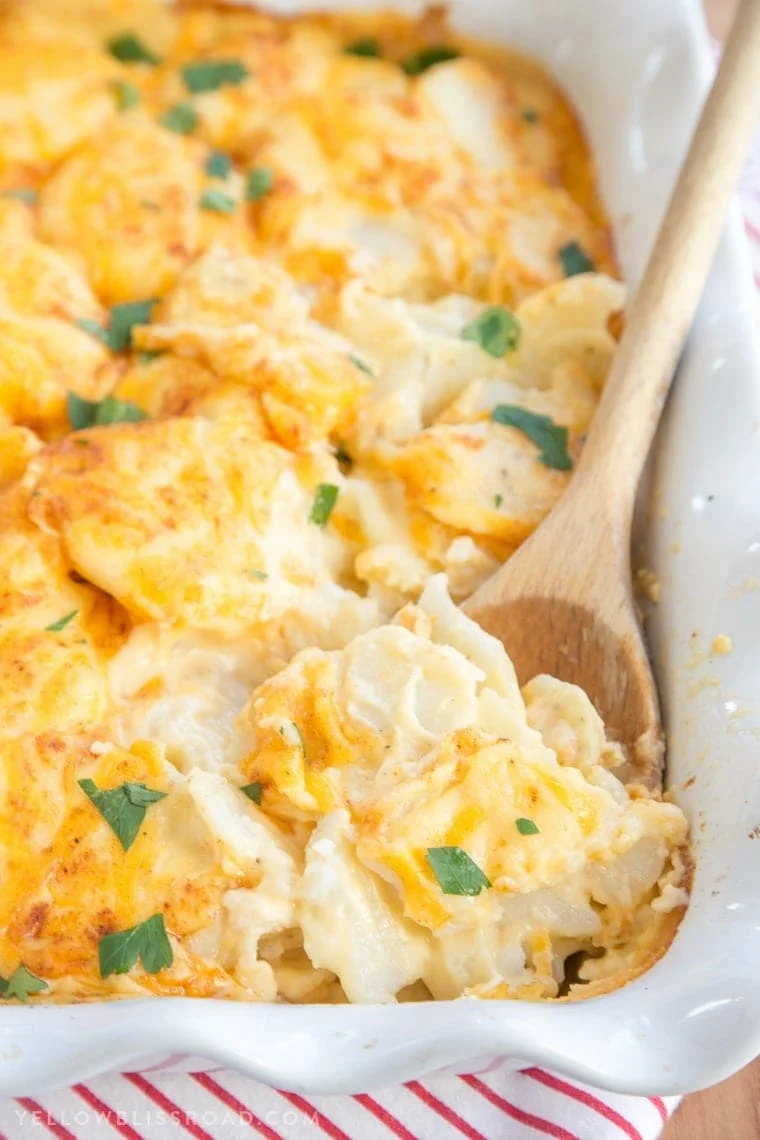 A baking dish filled with cheesy scalloped potatoes, topped with melted cheese and parsley, with a wooden spoon serving a portion—the perfect addition to your Easter side dishes.