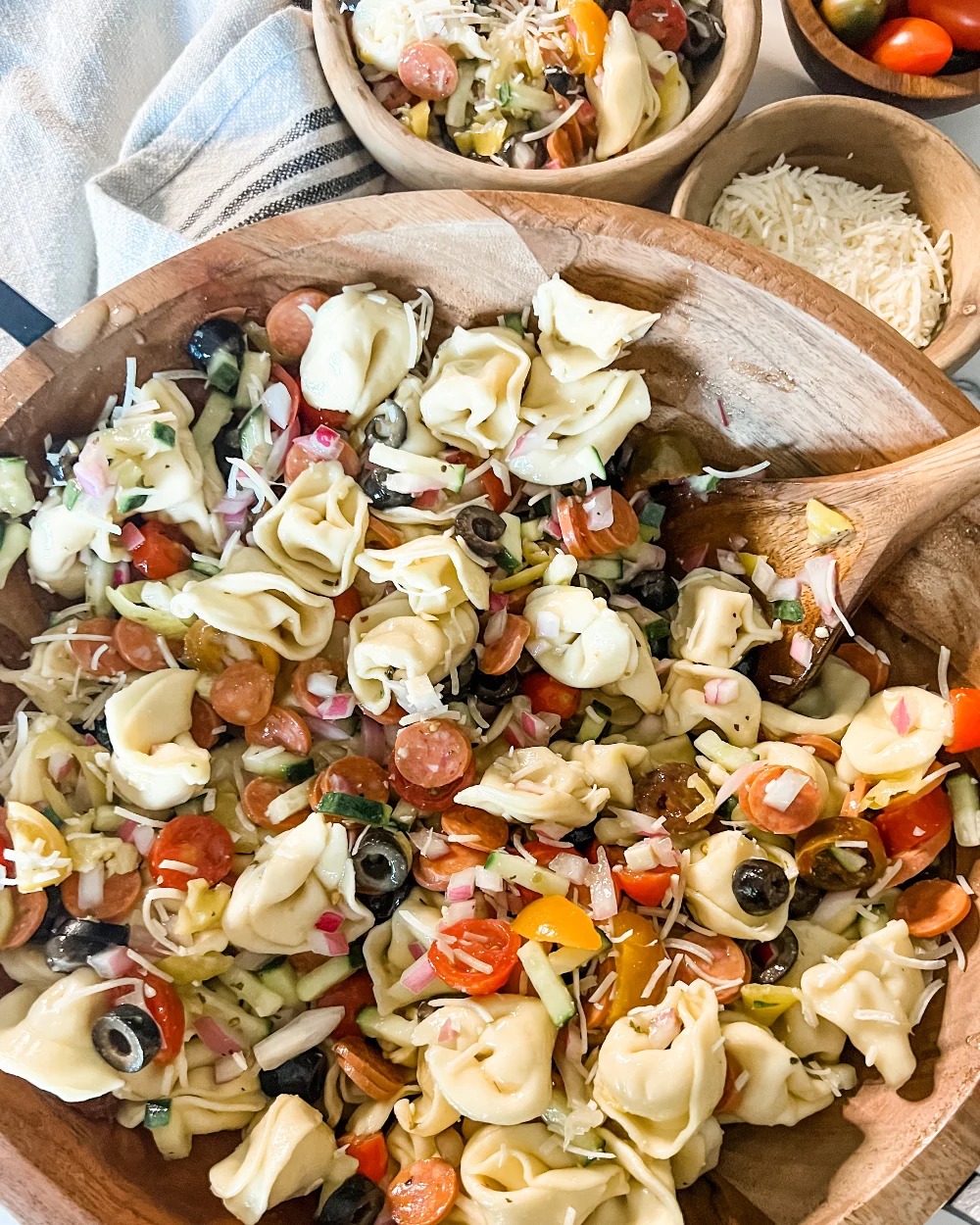 A large wooden bowl filled with tortellini pasta salad mixed with cherry tomatoes, olives, salami, onions, and cheese—perfect for Italian potluck ideas—with small bowls of salad and grated cheese nearby.