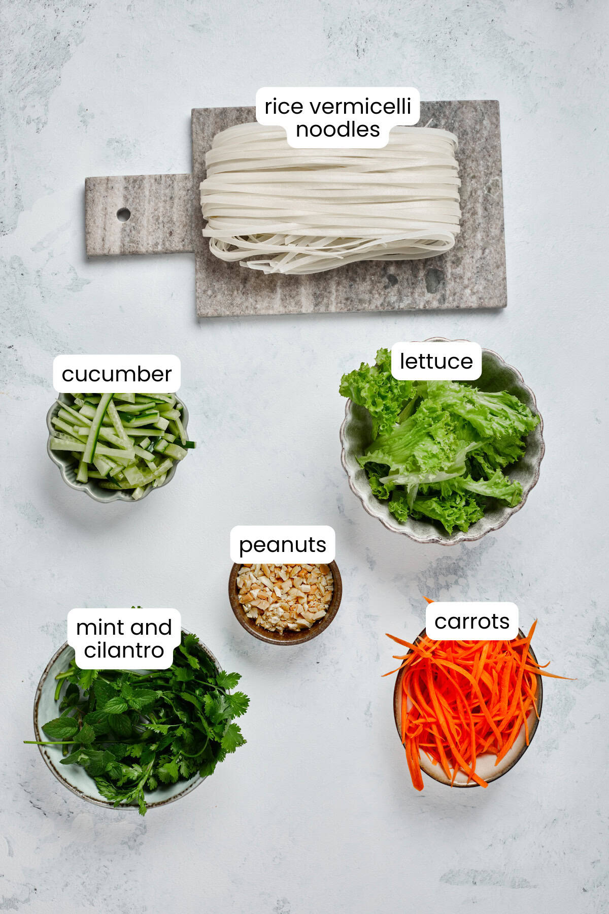 Ingredients for a Vietnamese Grilled Pork Rice Noodle Bowl are displayed separately: rice vermicelli noodles, cucumber, lettuce, peanuts, mint and cilantro, and carrots on a light background.