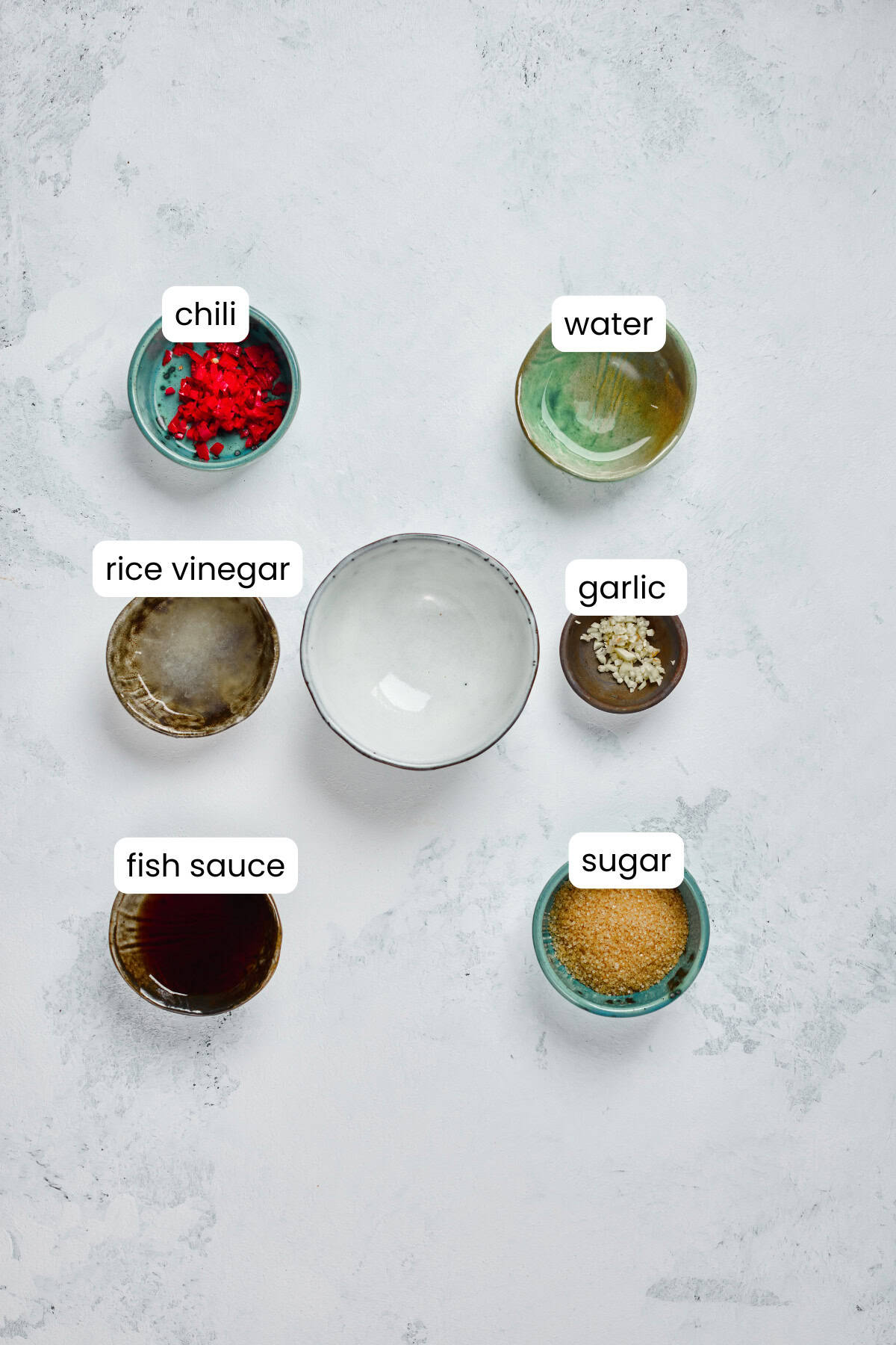 Six small bowls for a Vietnamese Noodle Bowl, holding chili, water, rice vinegar, garlic, fish sauce, and sugar, are arranged on a light surface. Each ingredient is labeled for easy prep of your Rice Noodle Bowl or Vietnamese Grilled Pork.