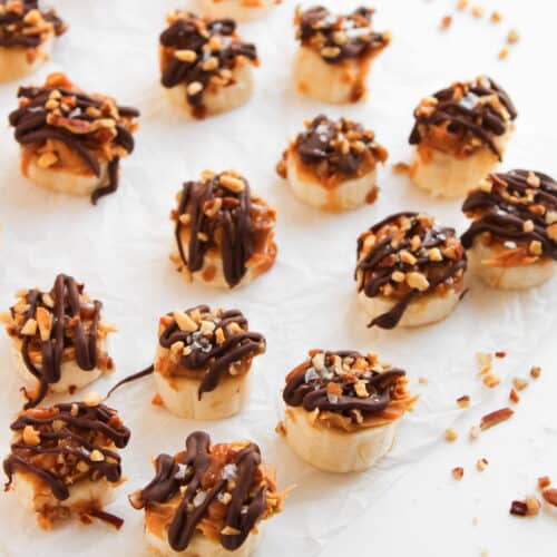 Bite-sized frozen banana slices topped with caramel, chopped nuts, and drizzled chocolate create irresistible Snickers Bites arranged on parchment paper.
