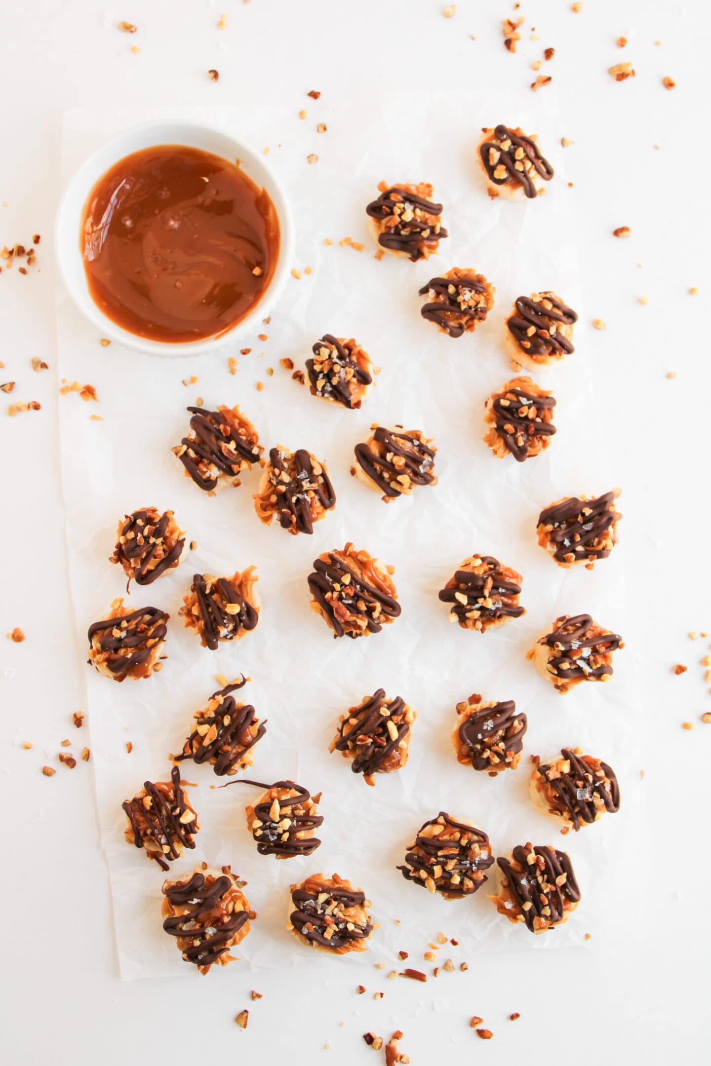 Overhead view of clusters of nut and chocolate-topped caramel candies on parchment paper, with a bowl of melted chocolate on the side—perfect for making your own Snickers Bites or Frozen Banana Snickers at home.