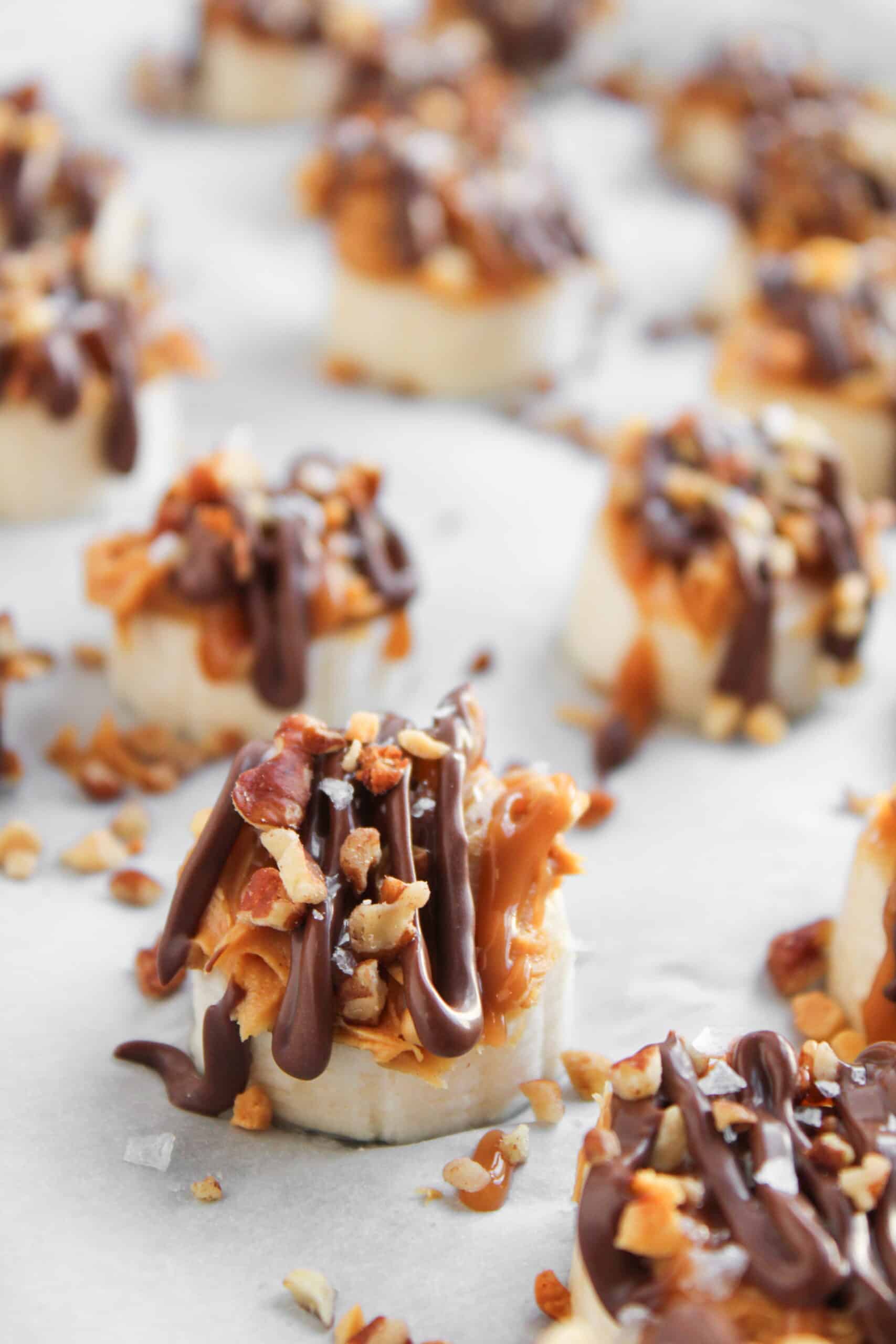 Frozen Banana Snickers Bites: Banana slices topped with caramel, chopped nuts, and drizzled with chocolate, arranged on parchment paper for a delicious frozen treat.