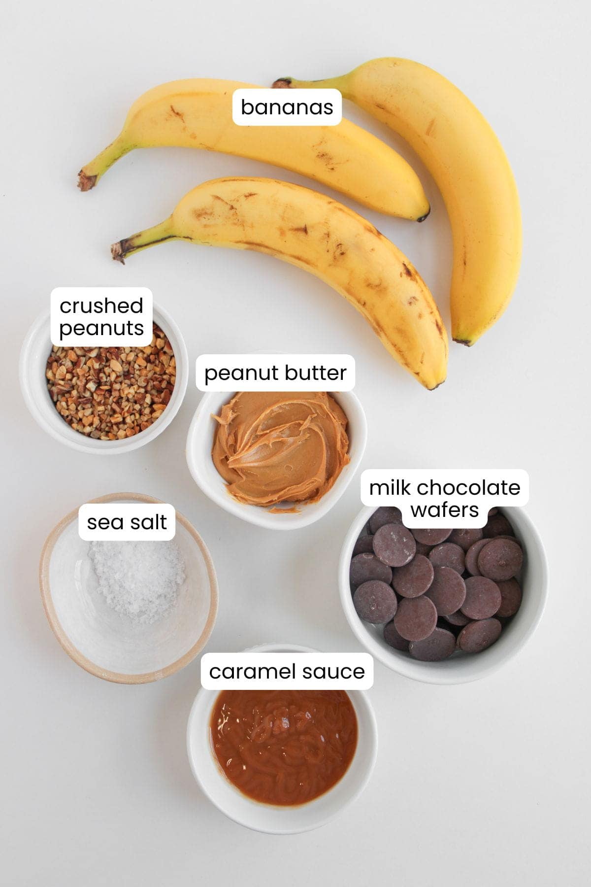 Three bananas, a bowl of crushed peanuts, peanut butter, milk chocolate wafers, sea salt, and caramel sauce are arranged on a white surface—perfect ingredients for making Frozen Banana Snickers Bites and other delicious frozen snacks.