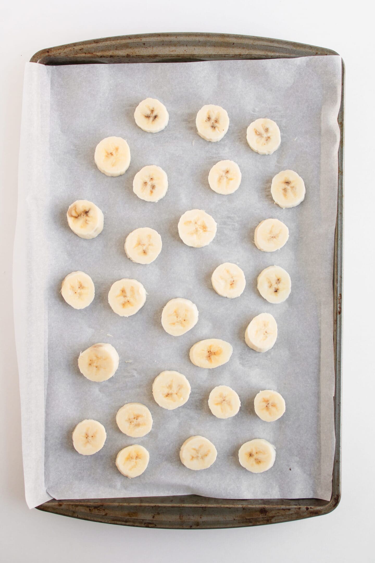 A baking sheet lined with parchment paper holds evenly spaced frozen banana slices arranged in rows, perfect for creating a delicious frozen dessert or Snickers bites.