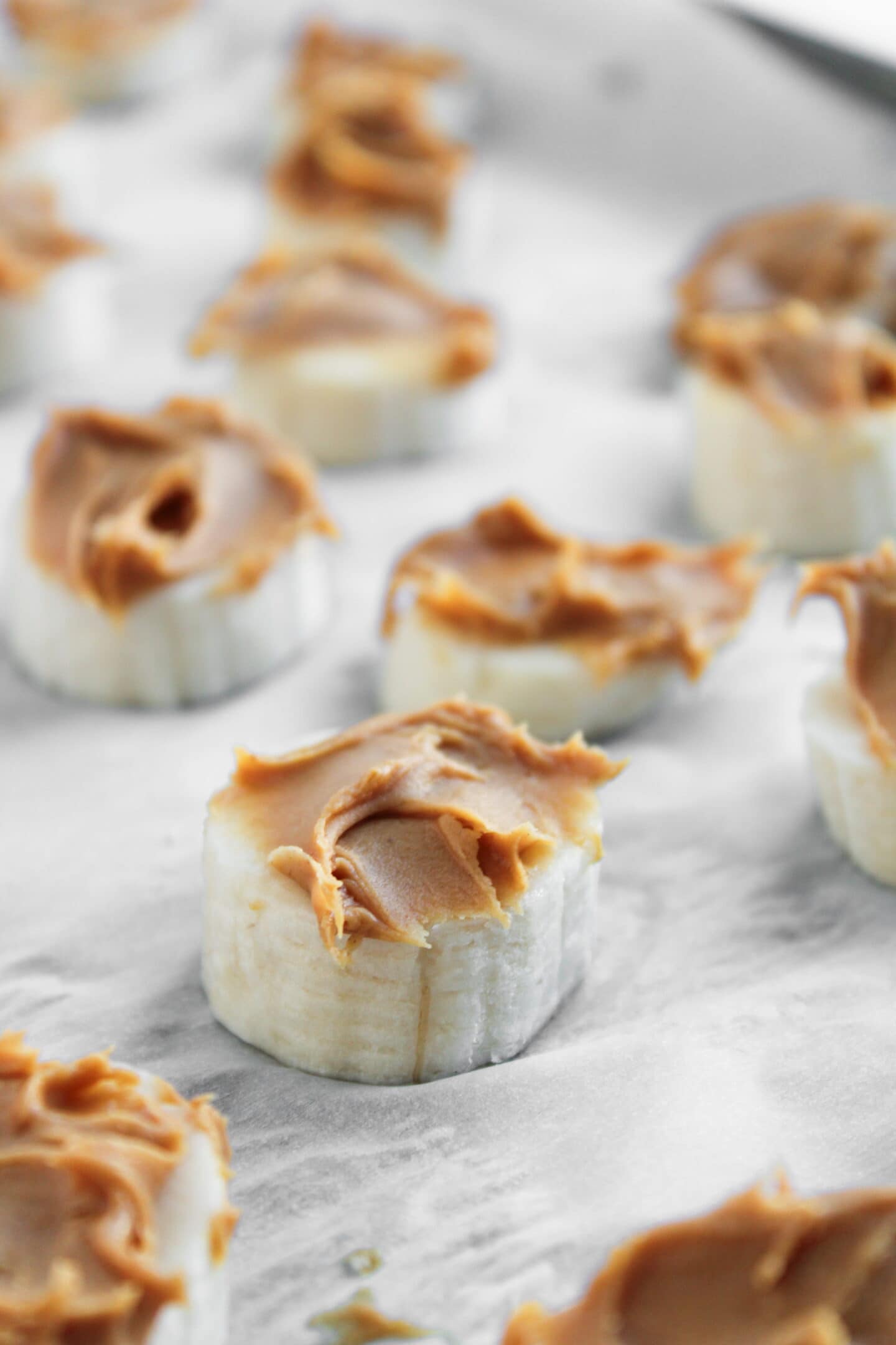 Sliced bananas topped with creamy peanut butter are arranged on a sheet of parchment paper for easy Frozen Banana Snickers snacks.