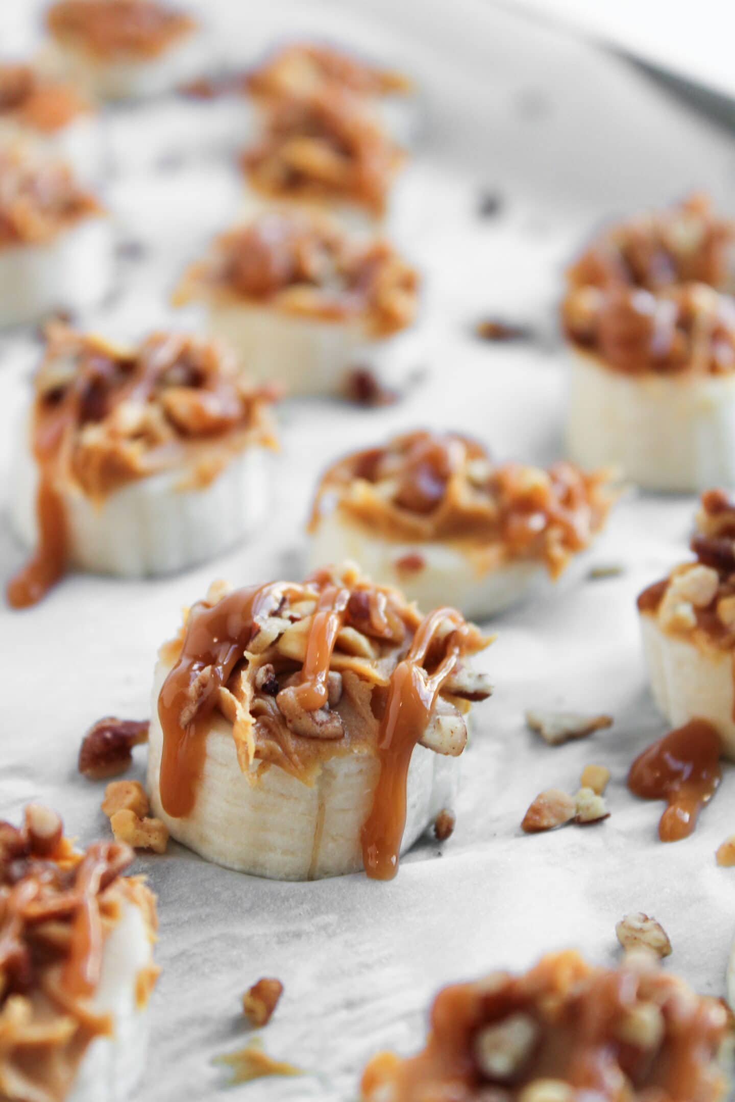 Frozen Banana Snickers slices topped with peanut butter, chopped nuts, and caramel drizzle arranged on a parchment-lined surface.