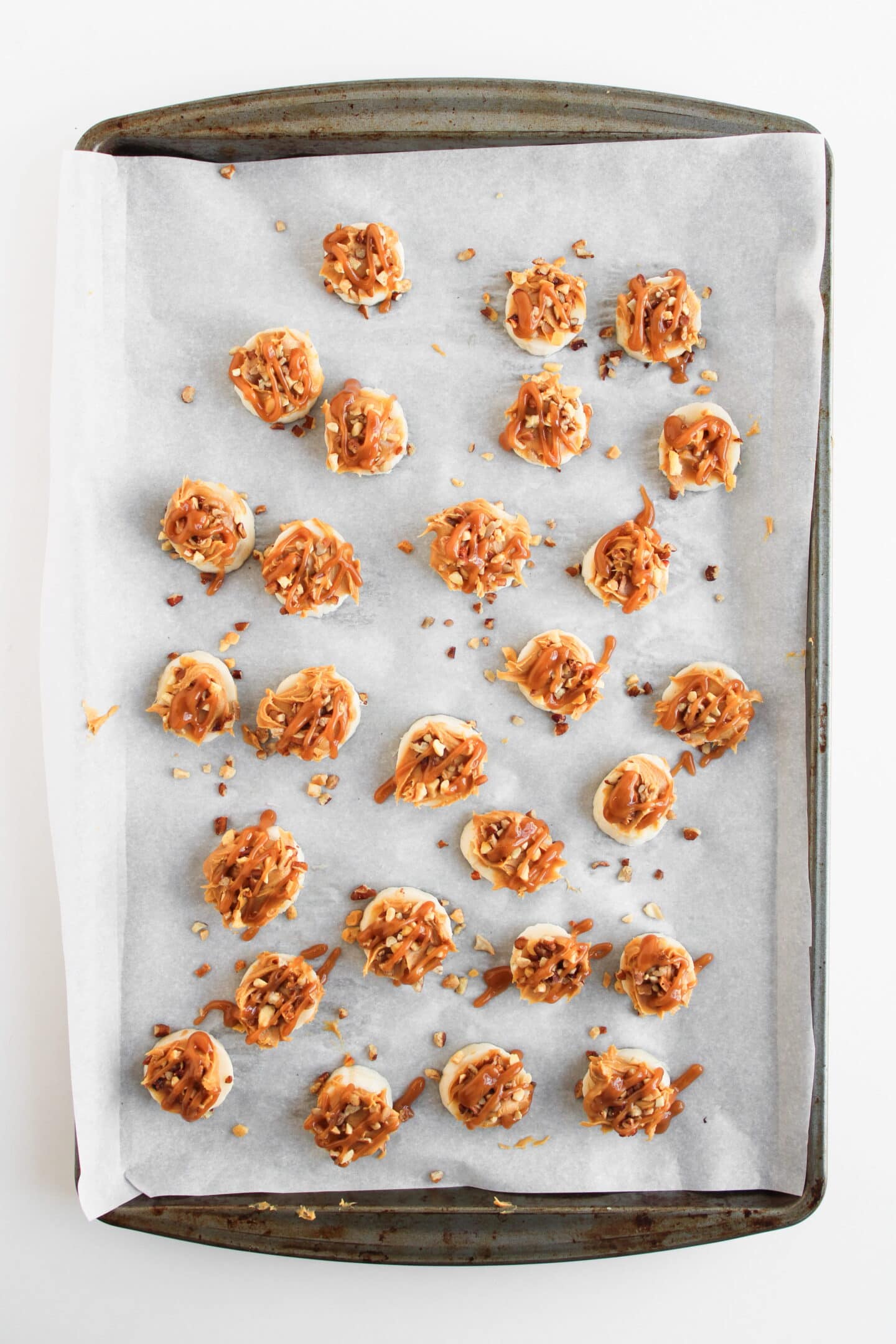 A baking sheet lined with parchment paper holds several round cookies topped with caramel drizzle, chopped nuts, and pieces of Snickers Bites, spaced out across the surface.