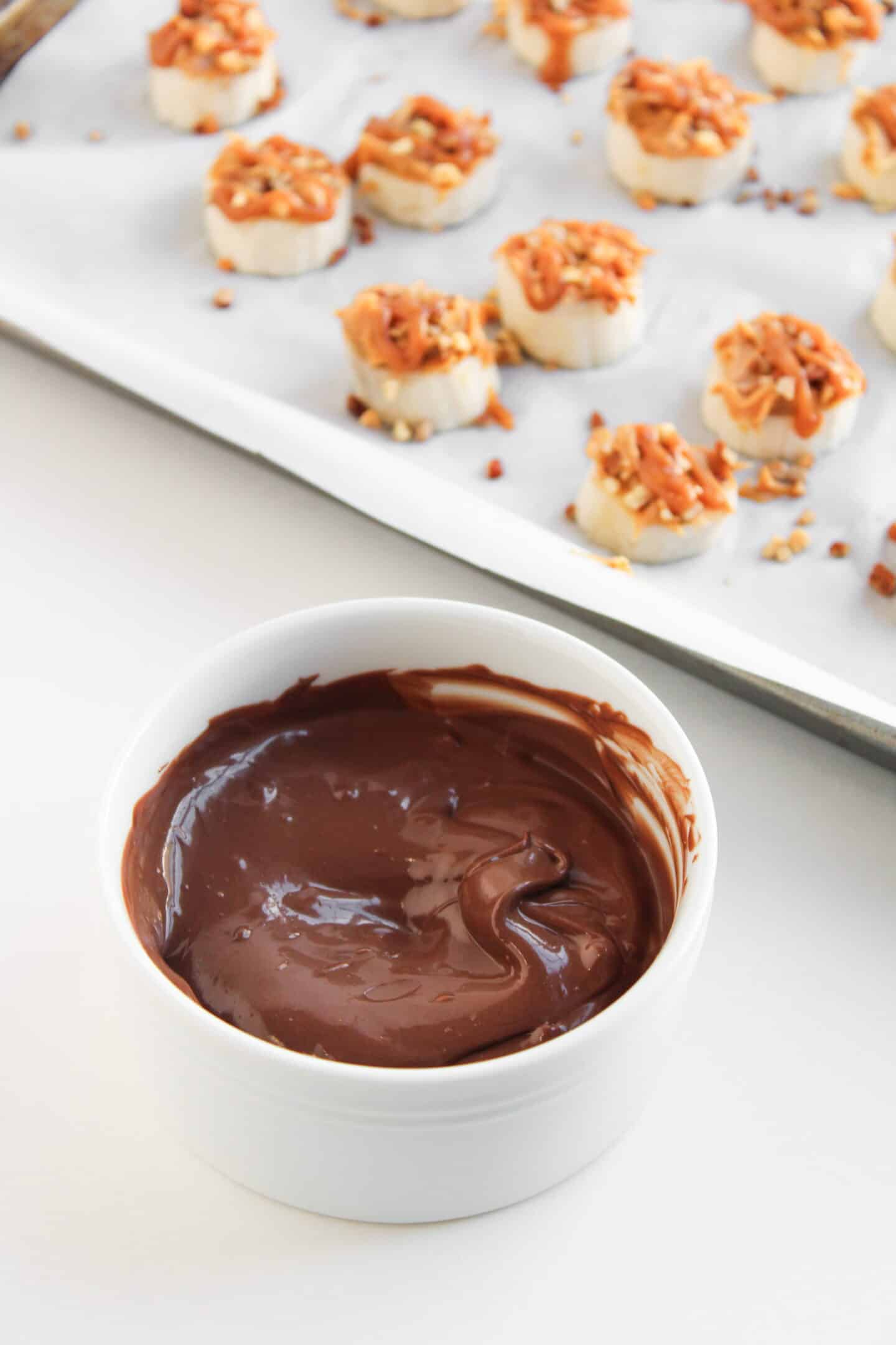 A white bowl of melted chocolate sits in front of a baking tray lined with Frozen Banana slices topped with nuts and caramel, perfect for making homemade Frozen Banana Snickers bites.