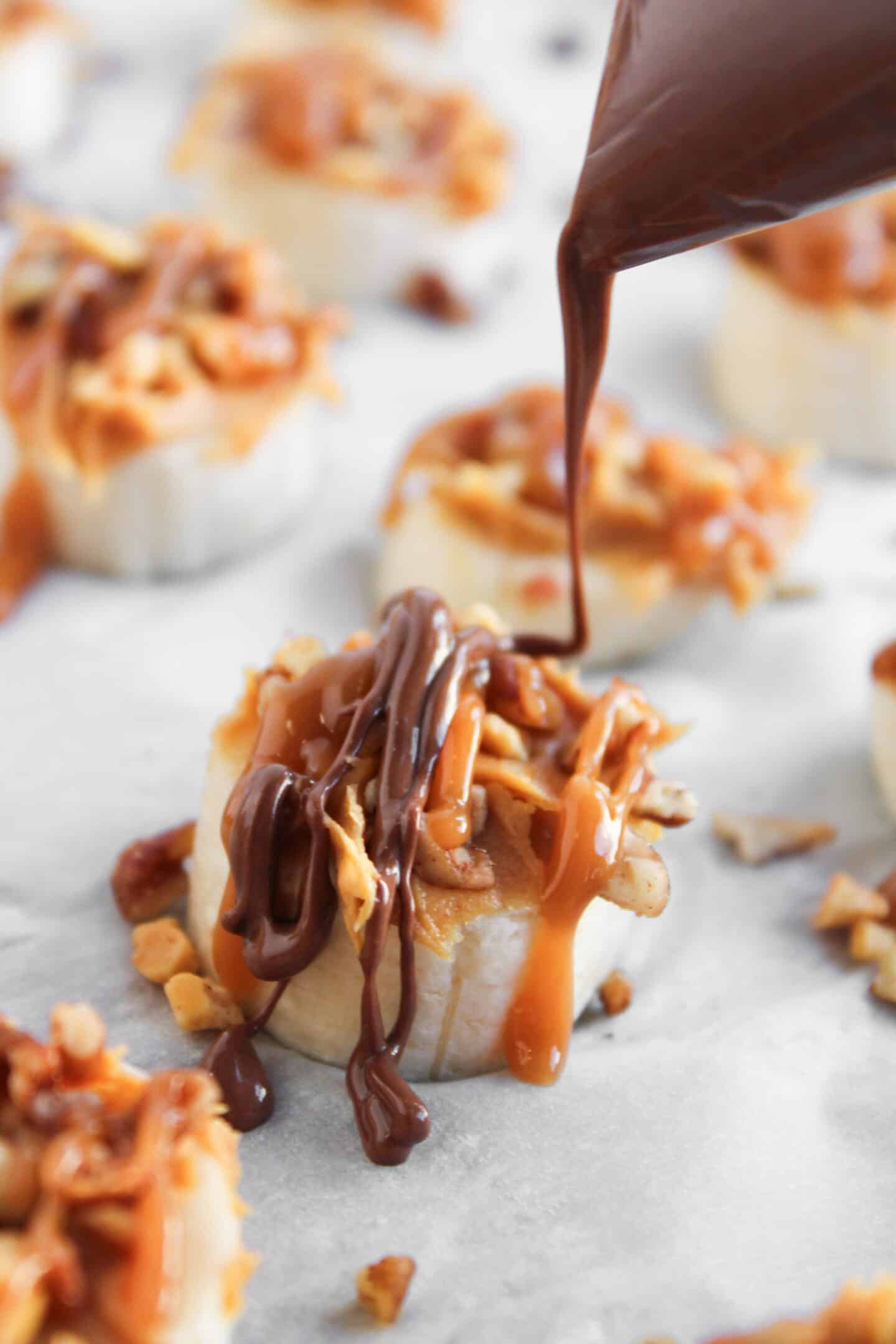 Frozen Banana Snickers Bites are made by topping sliced bananas with chopped nuts and caramel, then drizzling them with melted chocolate from a piping bag on parchment paper.