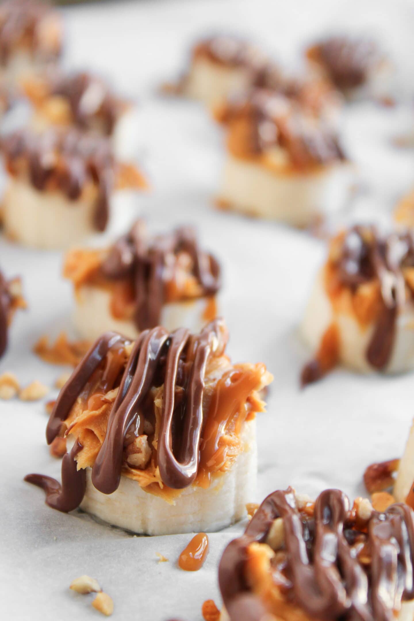 Sliced bananas topped with peanut butter, chopped nuts, and drizzled with chocolate and caramel on a parchment-lined surface—your easy homemade Frozen Banana Snickers treat.