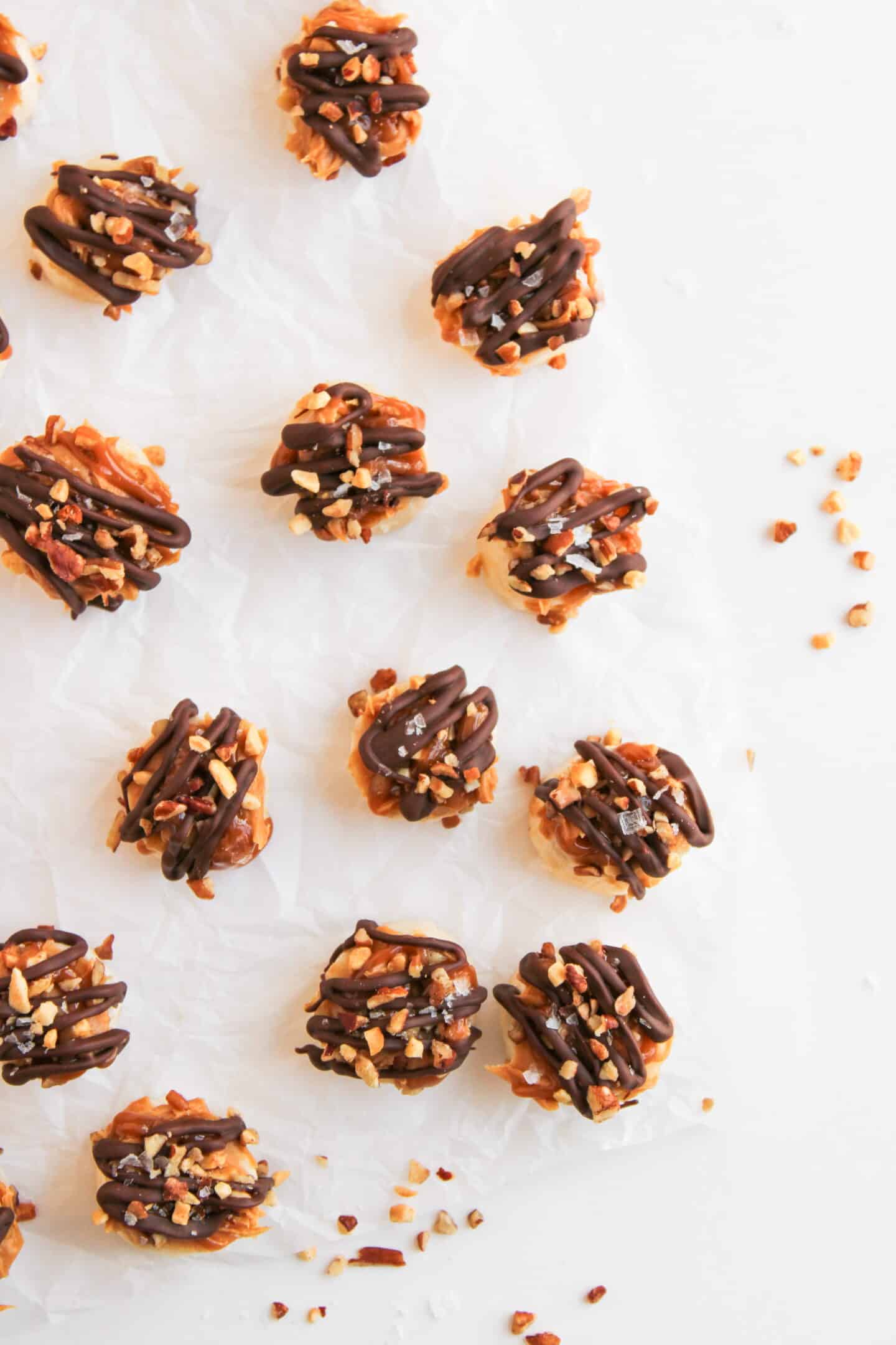 Small clusters of Frozen Banana Snickers are topped with chocolate drizzle and chopped nuts, arranged on parchment paper with scattered nut pieces around them on a white background.