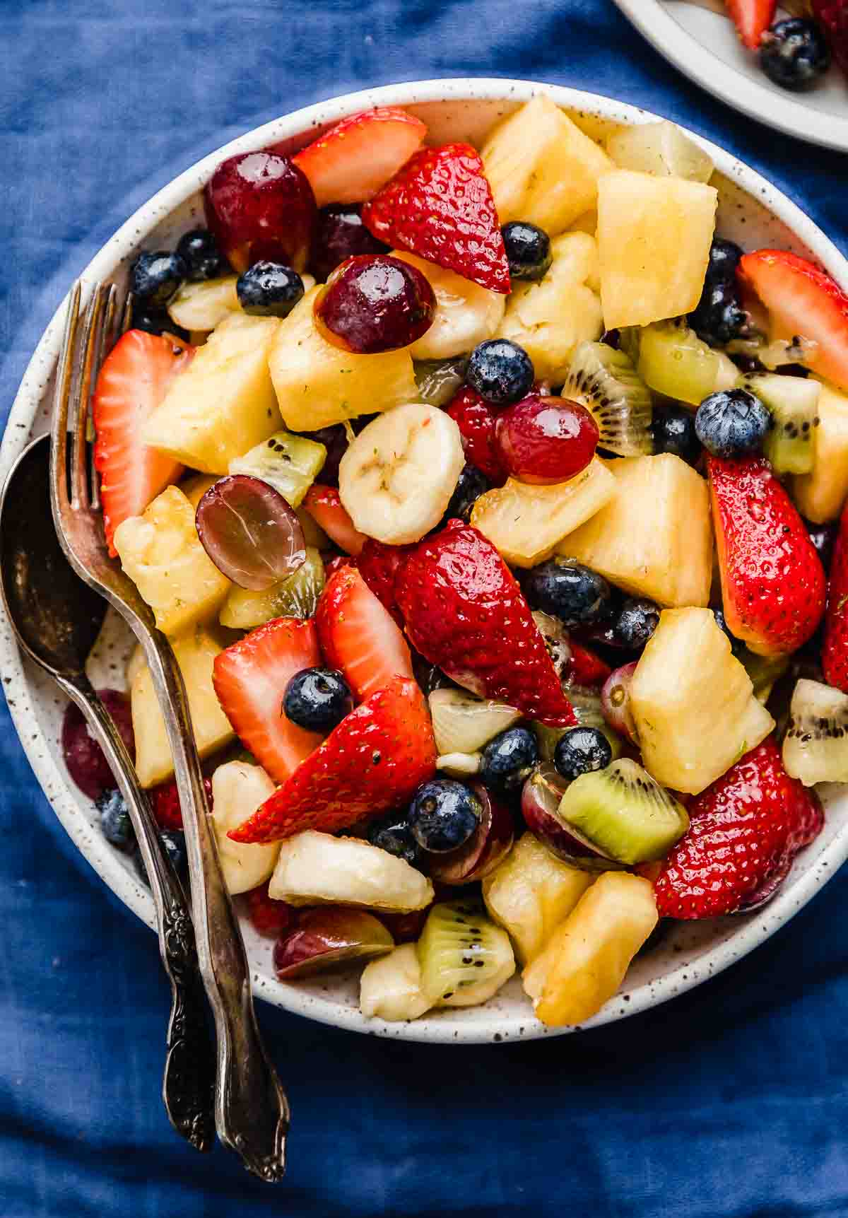A bowl of mixed fruit salad containing strawberries, blueberries, pineapple, banana, grapes, and kiwi sits with a fork and spoon on a blue surface—perfect as one of your refreshing Easter side dishes.