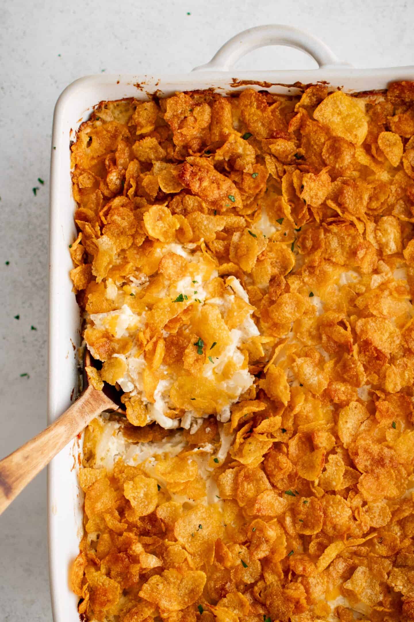 A close-up of a casserole dish filled with a cheesy, creamy baked dish topped with golden cornflakes—perfect for serving as one of your Easter side dishes. A serving spoon is scooping out a portion.