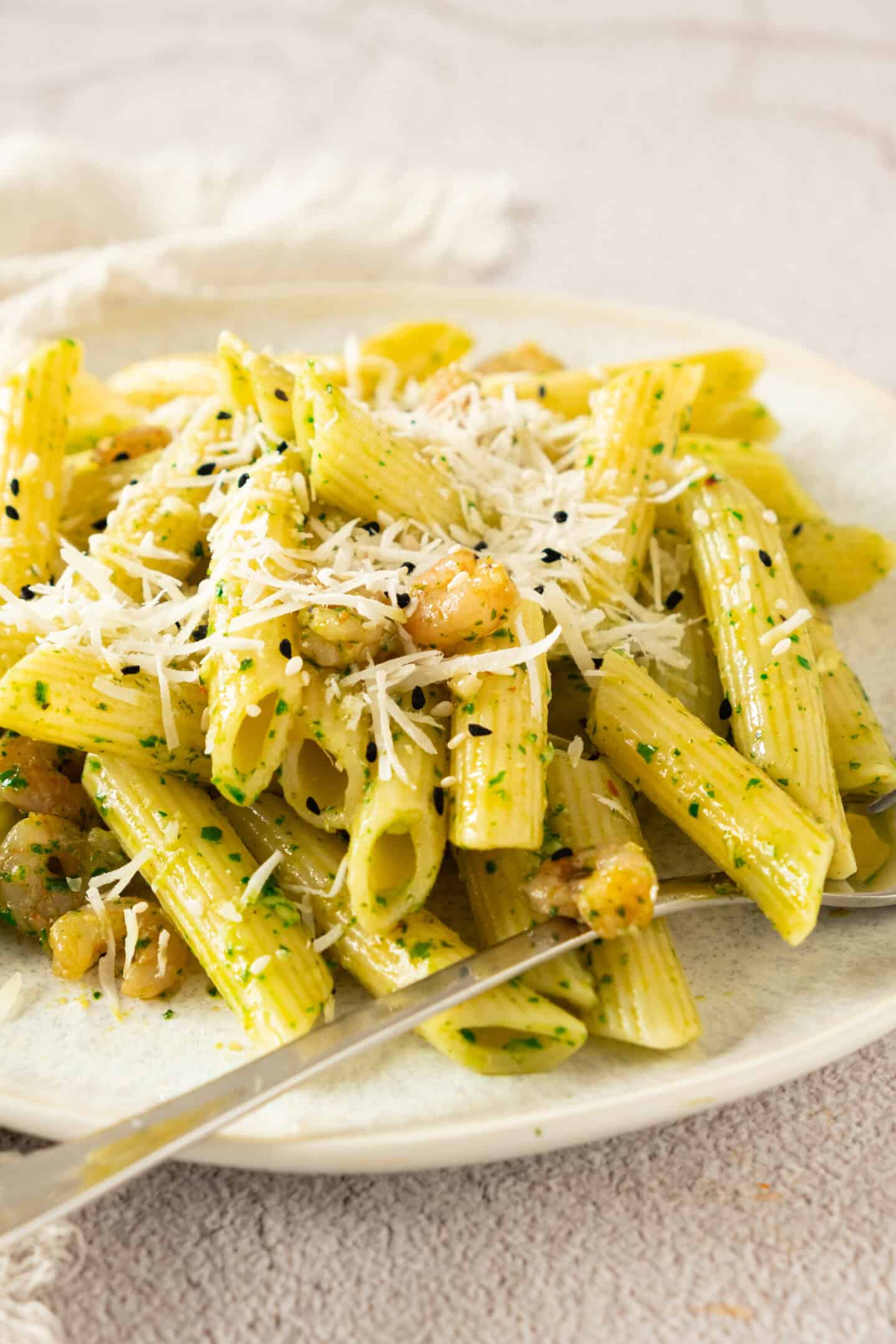 A plate of penne pasta mixed with pesto sauce, shrimp, and topped with grated cheese, served with a fork.