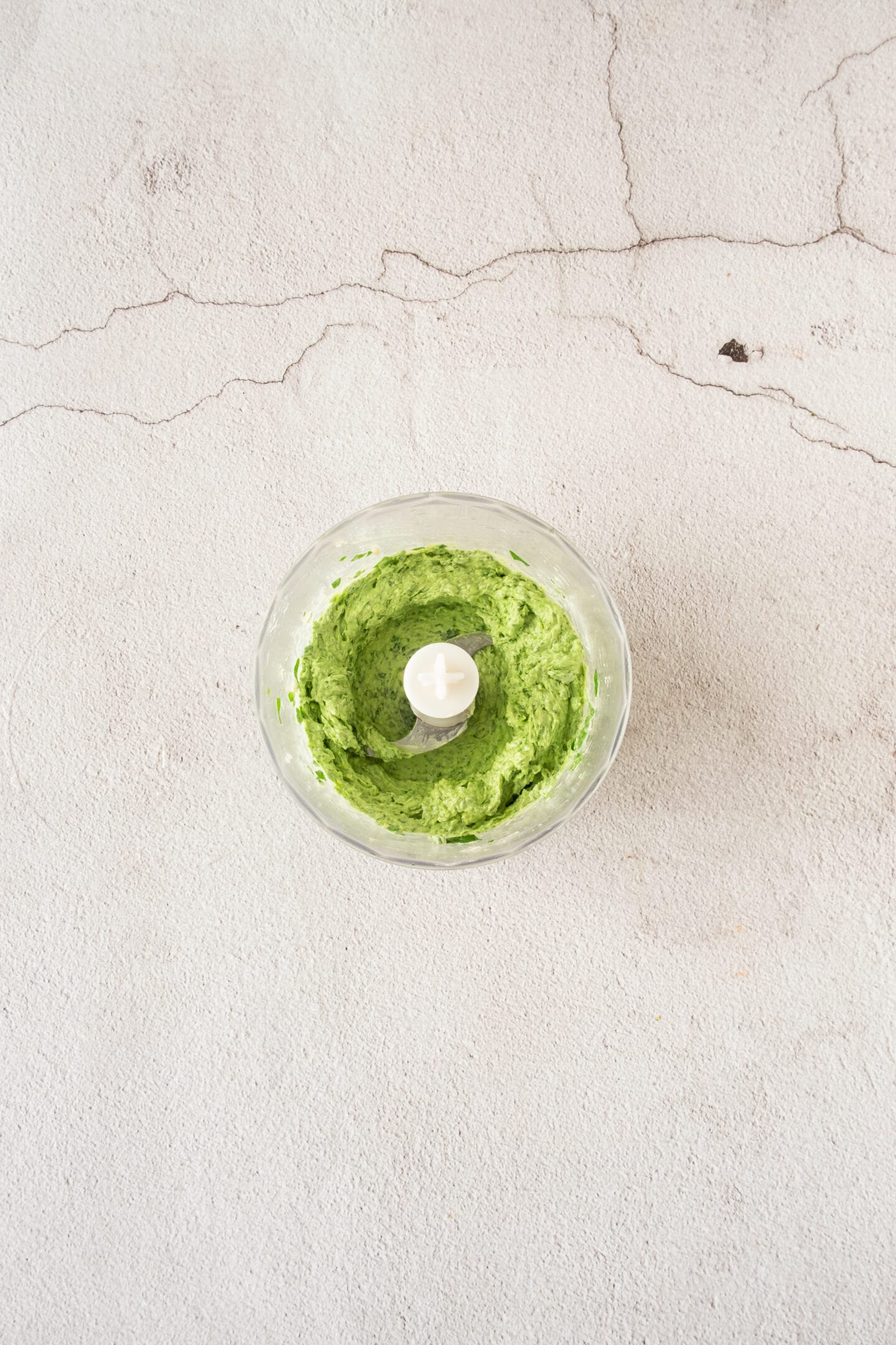 A food processor bowl containing green pesto sits on a light textured surface.
