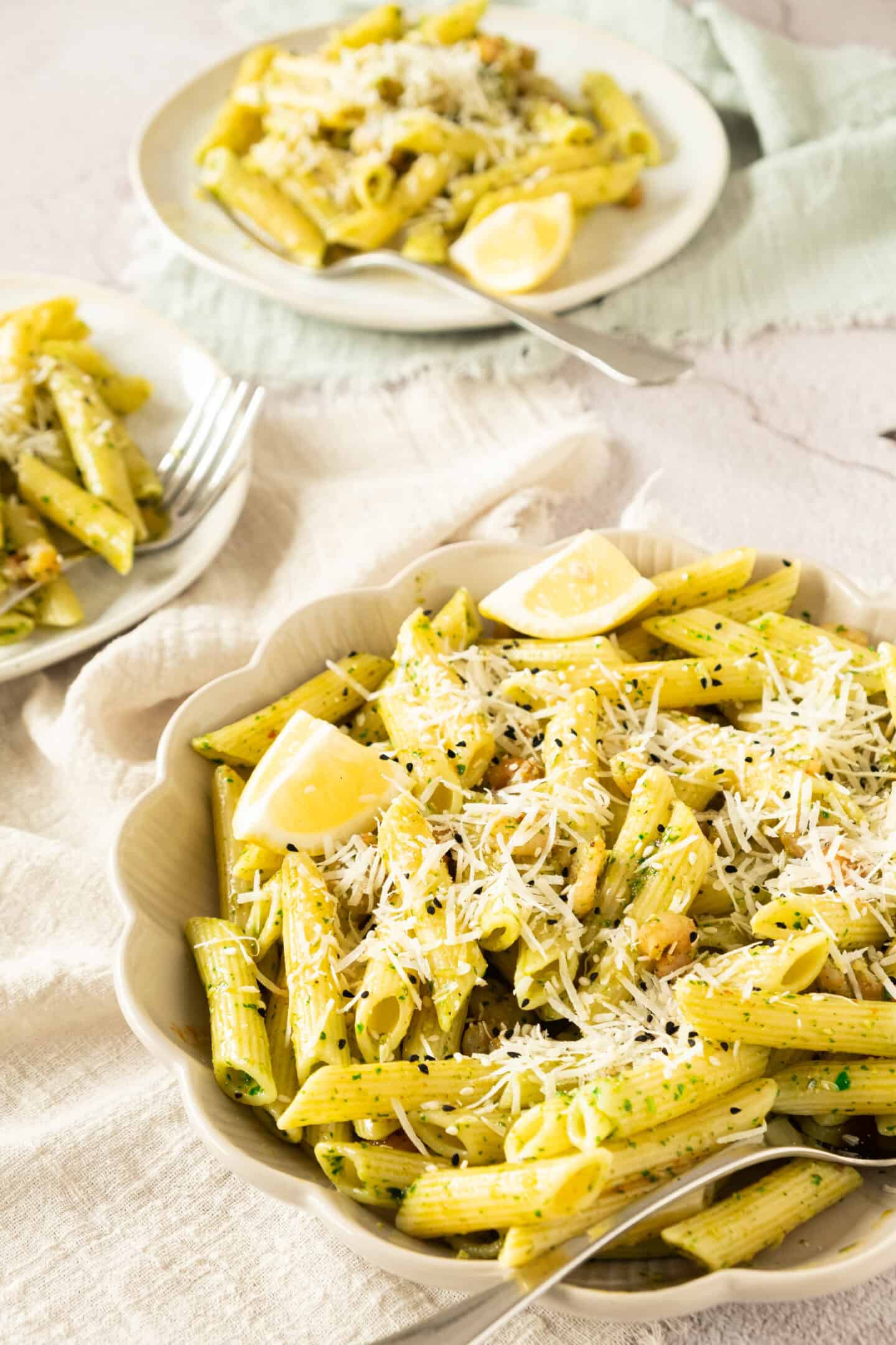 A bowl of penne pasta with pesto sauce, grated cheese, and lemon wedges, with two plates of pasta in the background.