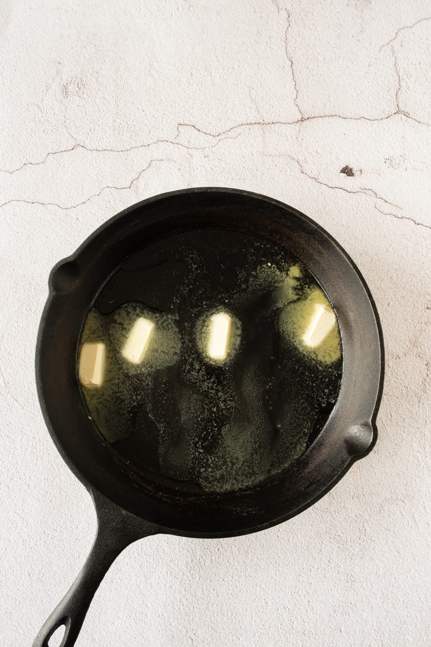 A cast iron skillet with several pieces of butter melting inside, placed on a light-colored, textured surface.