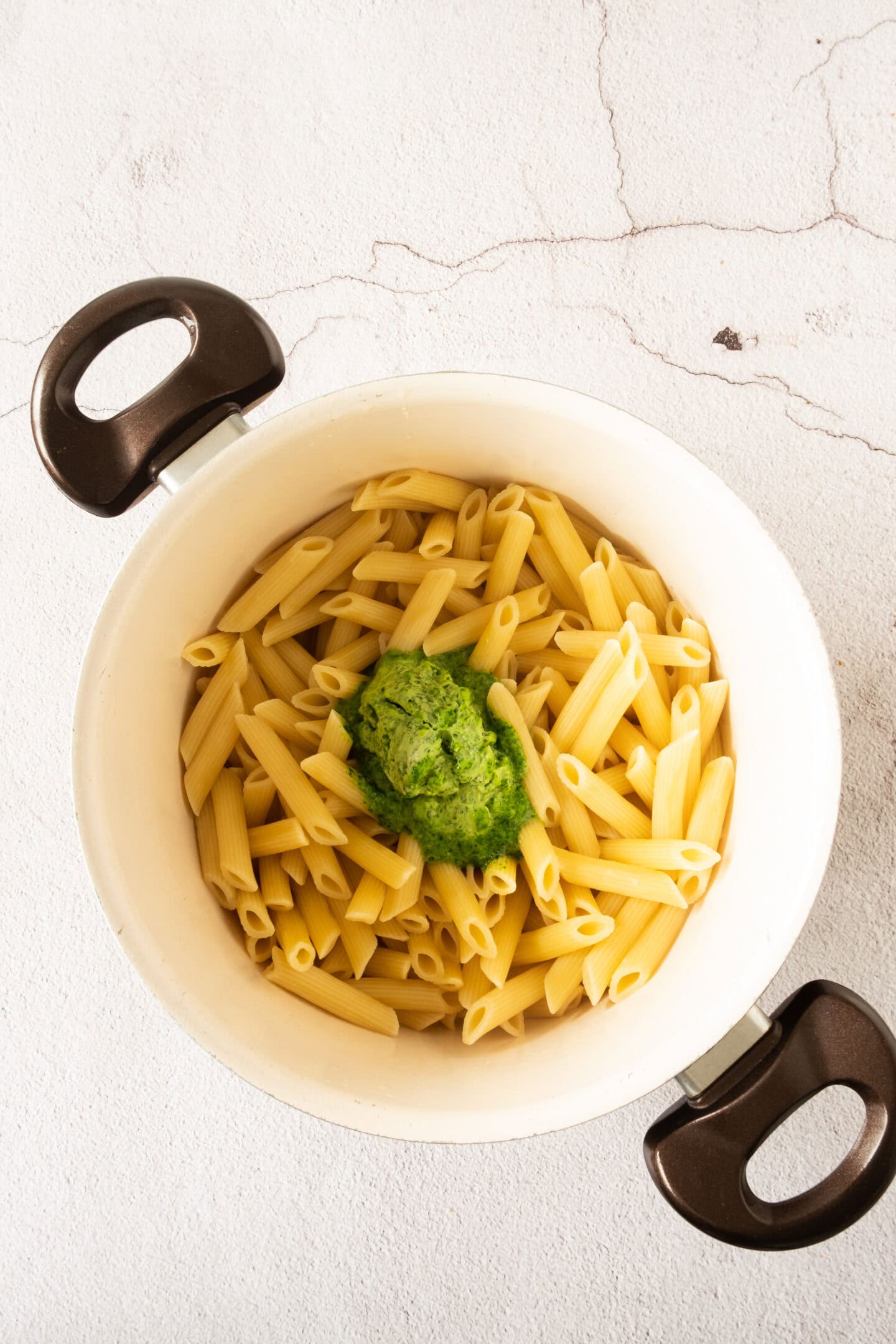A white pot containing cooked penne pasta with a scoop of green pesto sauce on top, placed on a light-colored surface.