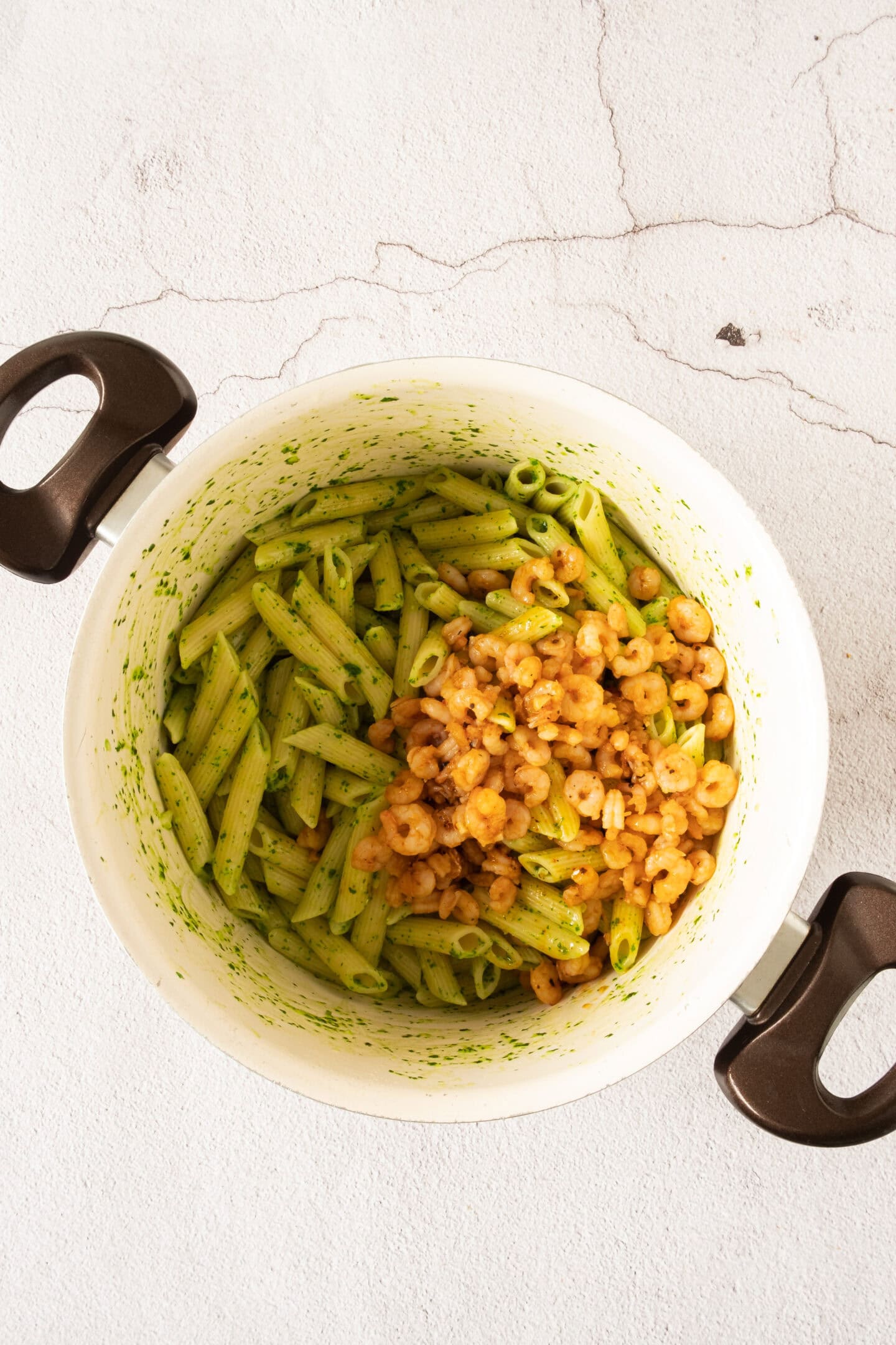 A white pot with handles contains green pesto penne pasta and cooked shrimp, placed on a light textured surface.