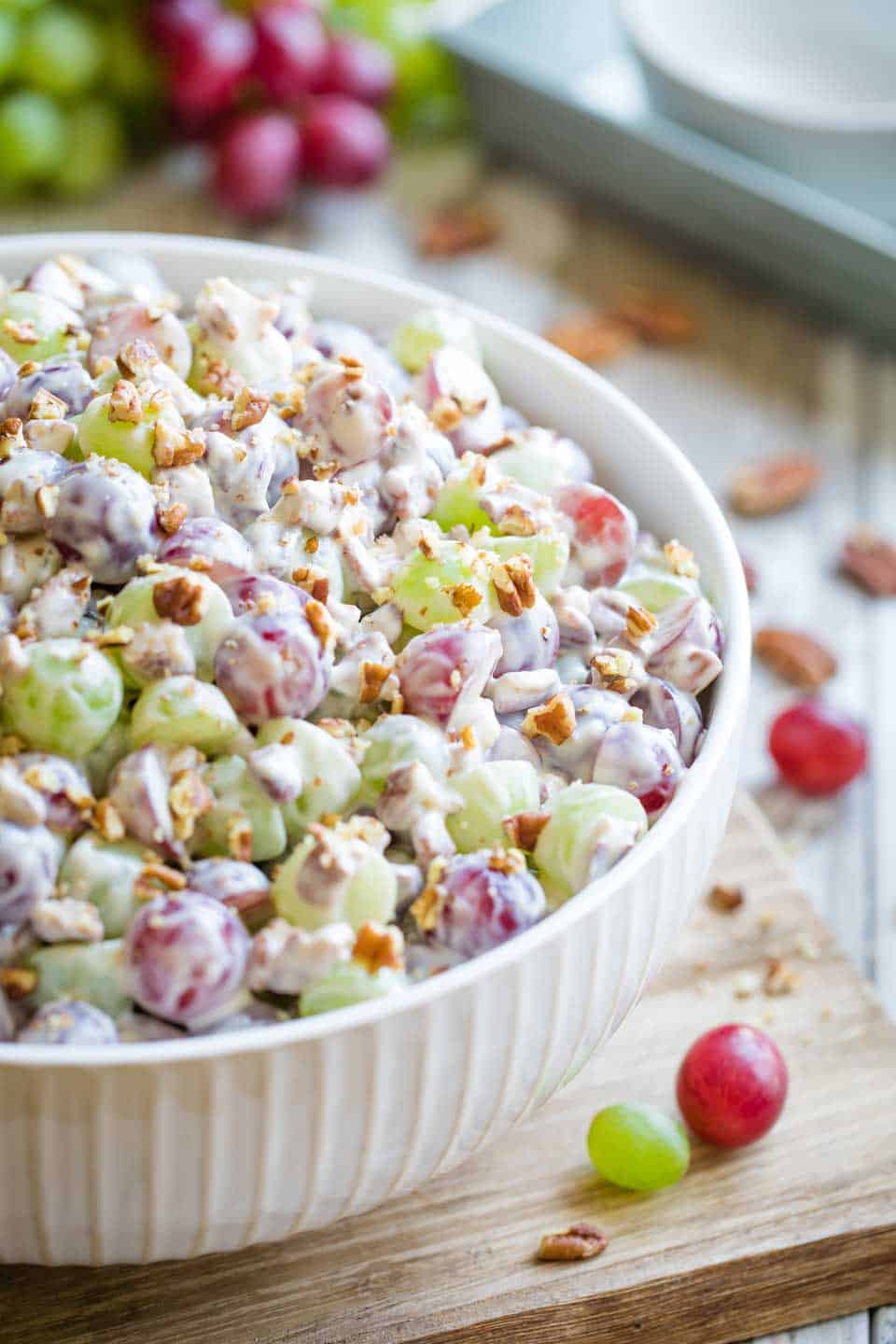 A white bowl filled with creamy grape salad topped with chopped pecans sits on a wooden surface, making it a delightful addition to your Easter side dishes, with fresh grapes visible in the background.