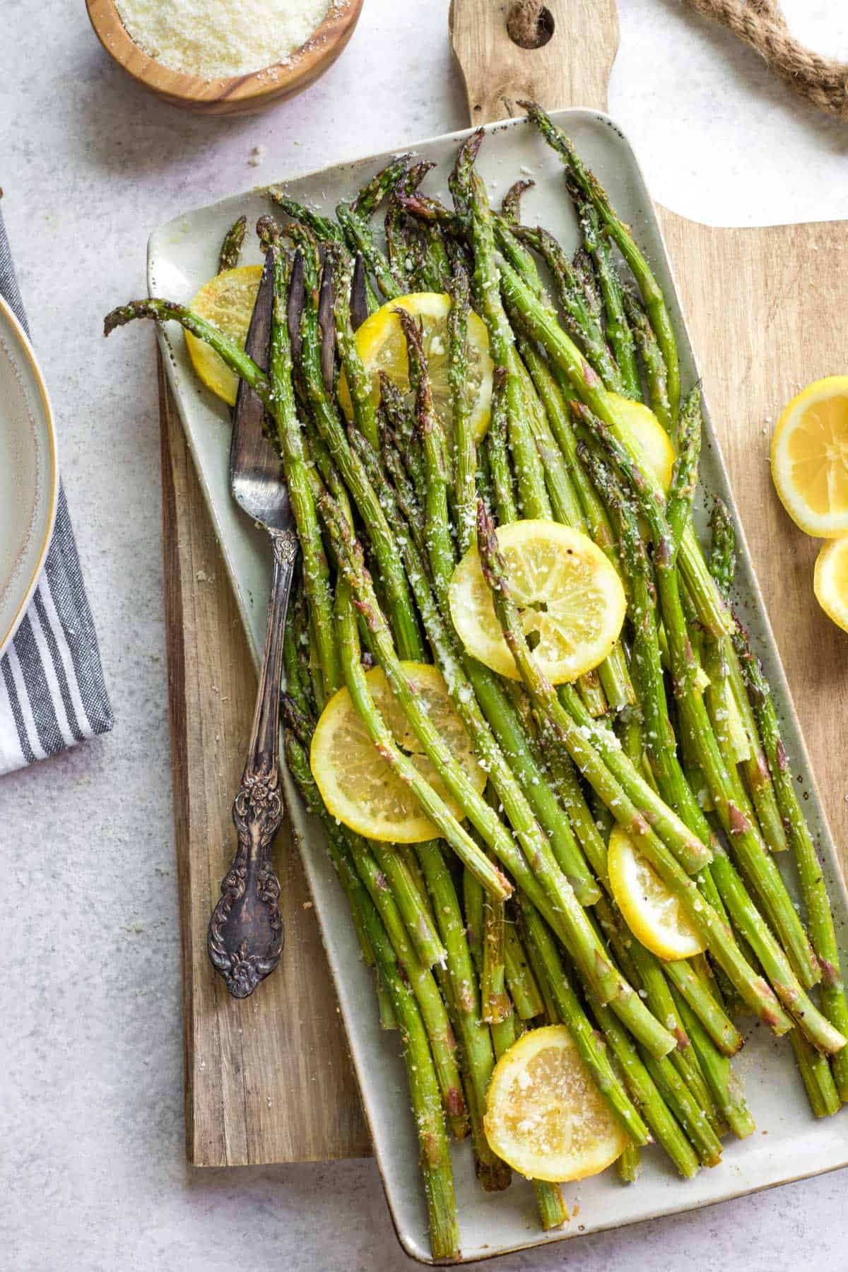 Roasted asparagus spears on a rectangular platter, garnished with lemon slices and grated cheese—an elegant touch for your Easter side dishes, served with a vintage fork and a half lemon nearby.