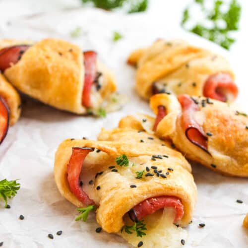 Golden-brown Ham and Cheese Rolls made with crescent rolls, filled with savory ham and melted cheese, sprinkled with black sesame seeds and garnished with fresh parsley on parchment paper.