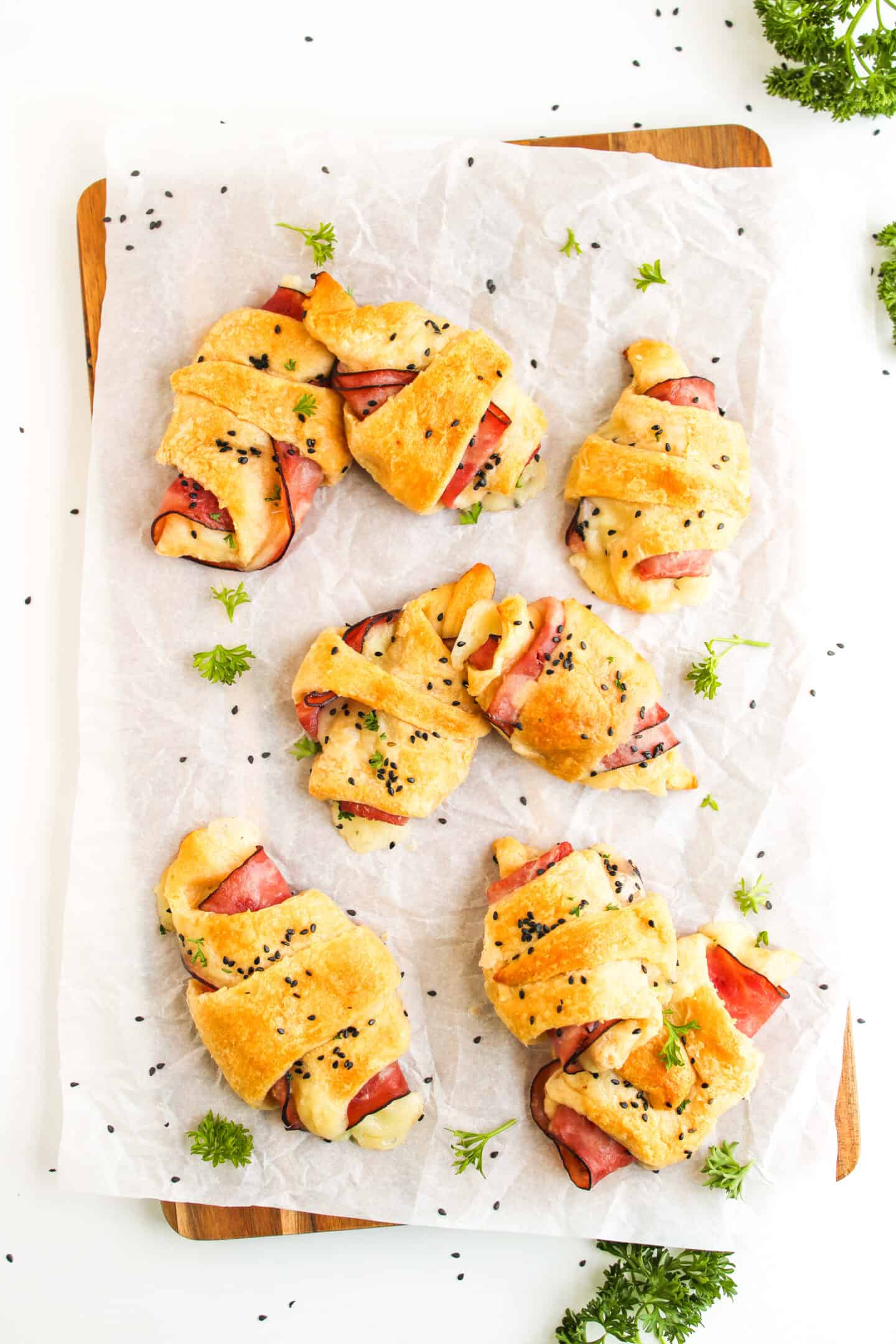 Six Ham and Cheese Crescent Rolls topped with black sesame seeds are arranged on parchment paper over a wooden board, garnished with fresh parsley.