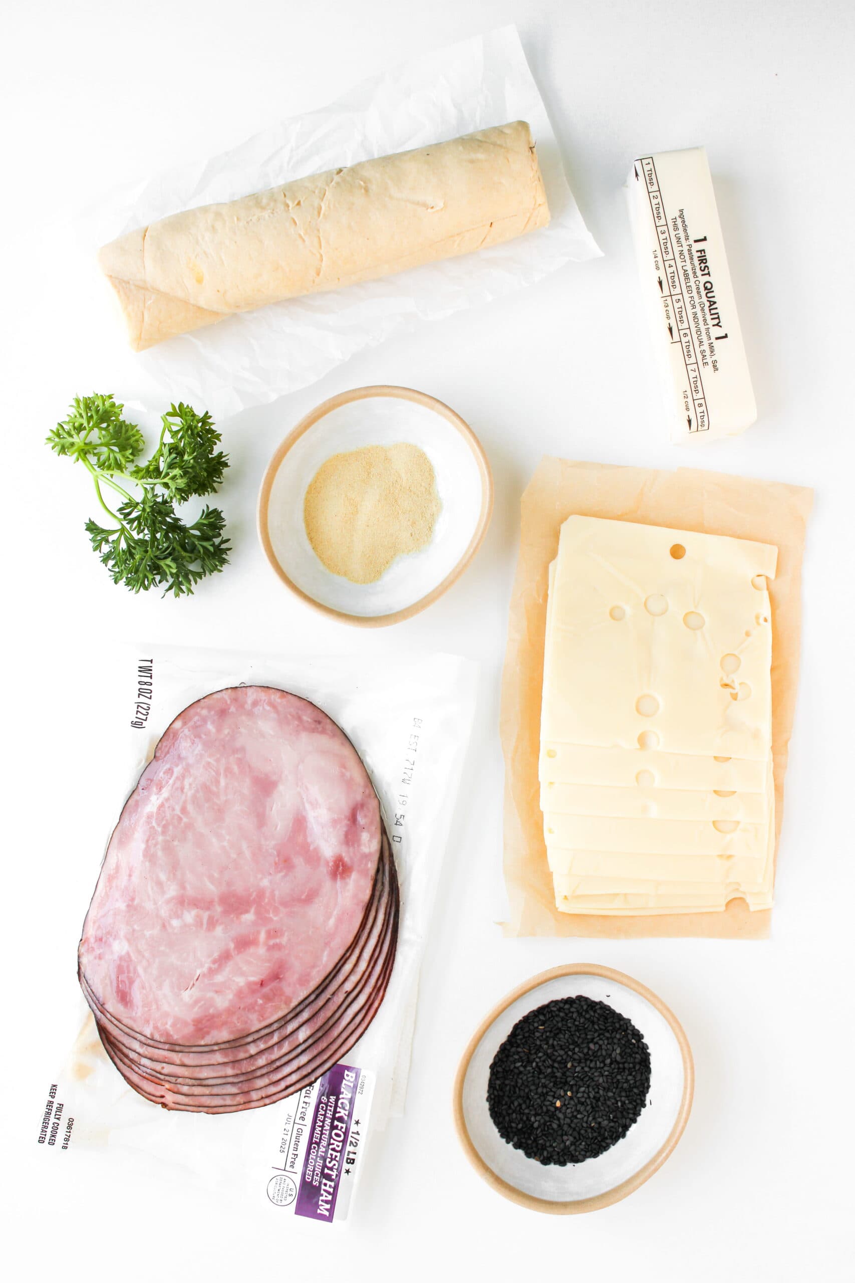 A baguette, butter, parsley, garlic powder, Swiss cheese slices, ham—perfect ingredients for Ham and Cheese Crescent Rolls—plus a small bowl of black sesame seeds arranged on a white surface.