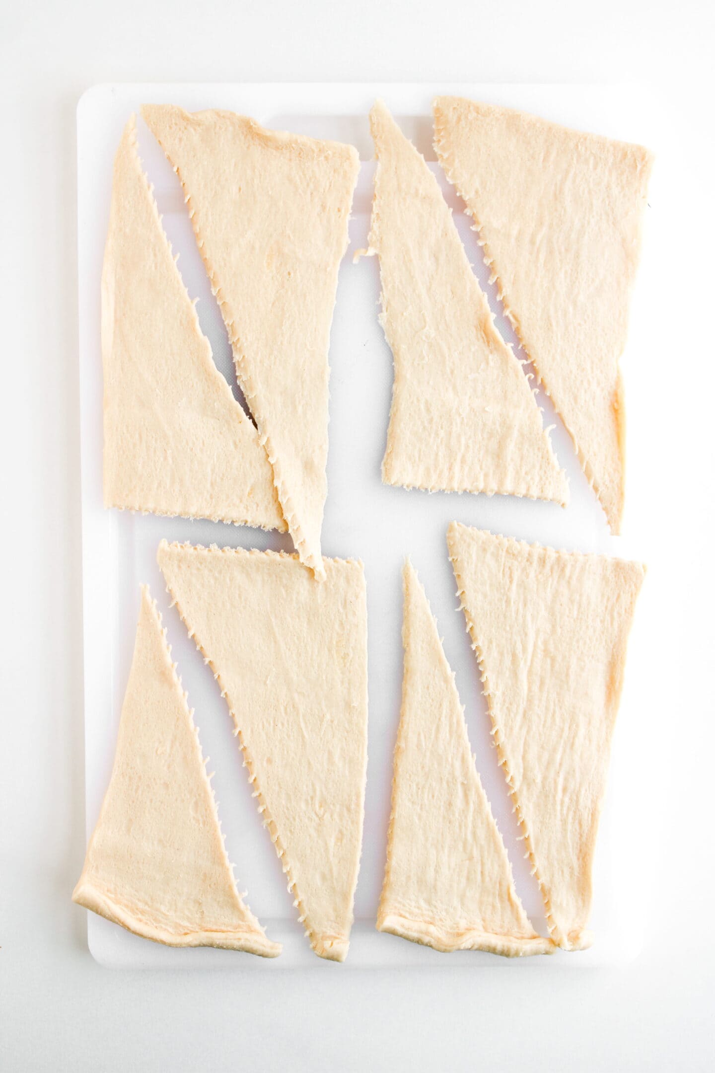 Rectangular sheet of raw dough cut into eight triangular pieces, ready to be rolled up into delicious Ham and Cheese Crescent Rolls, arranged on a white cutting board.